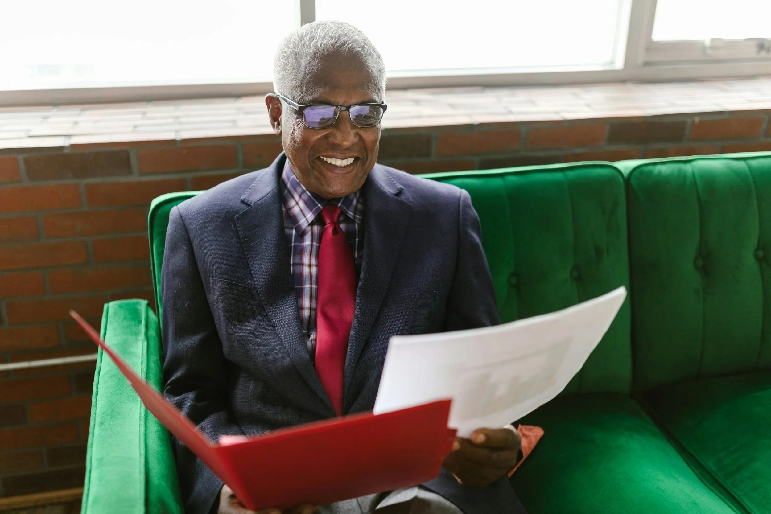 black senior university president sitting on a green couch wearing a suit and tie holding a red folding looking at paperwork and smiling