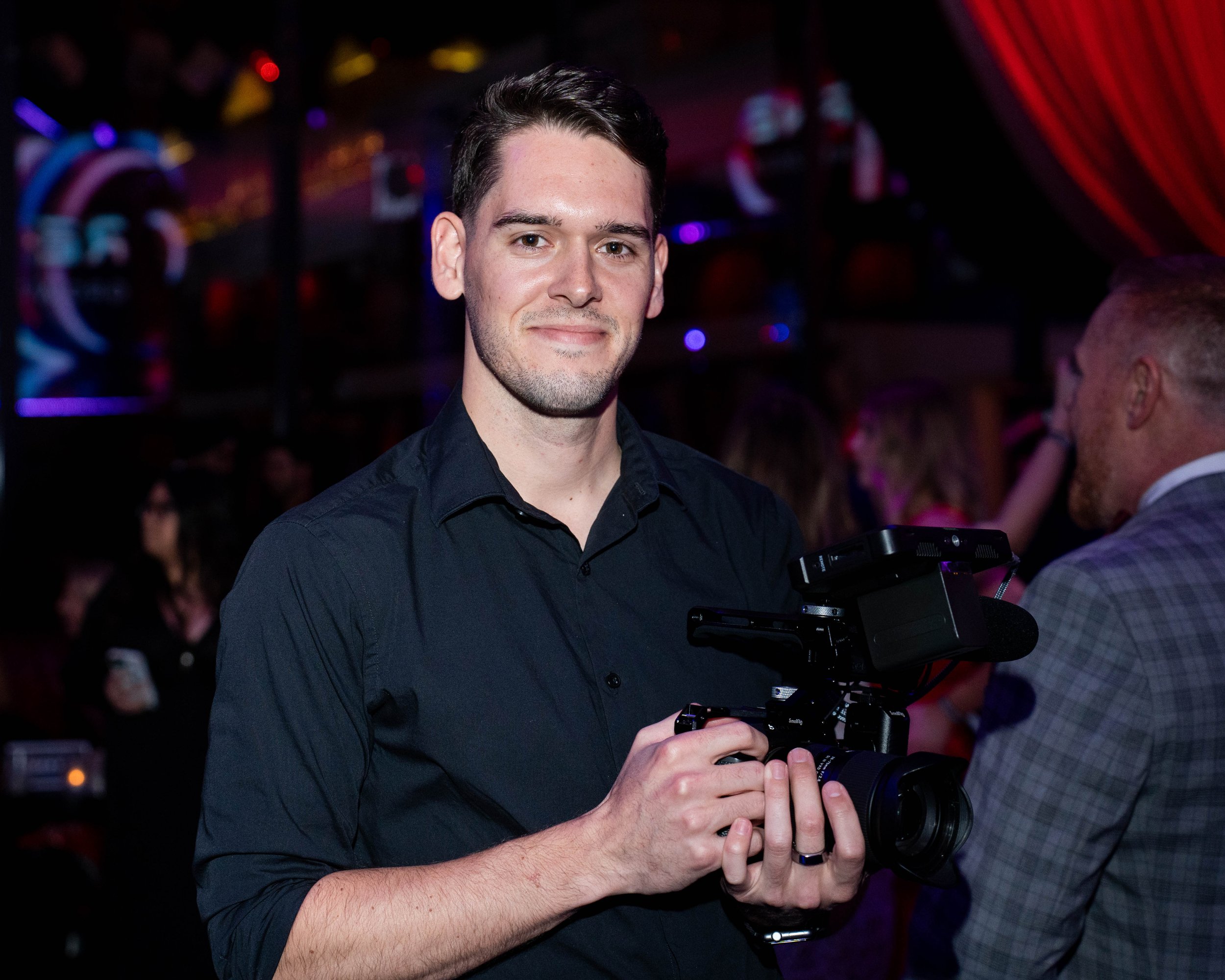 A man in a black shirt holding a professional video camera at a lively event or party.