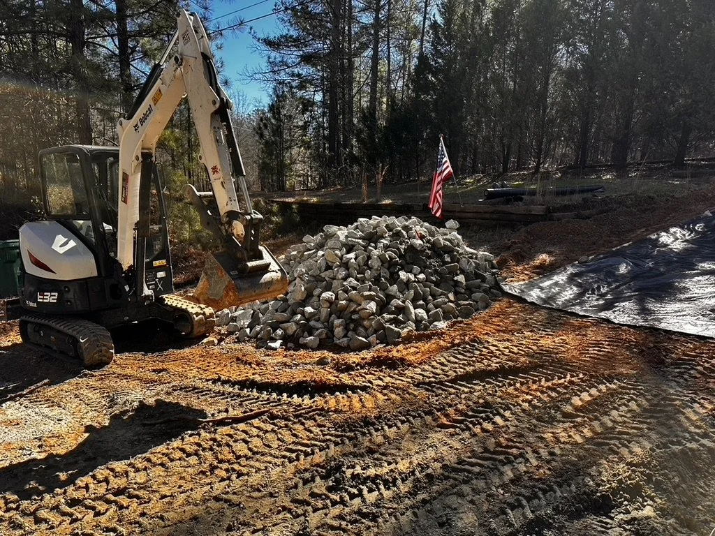 Construction site with a small excavator beside a pile of rocks, an American flag, and tracks on dirt ground surrounded by trees and a black tarp.