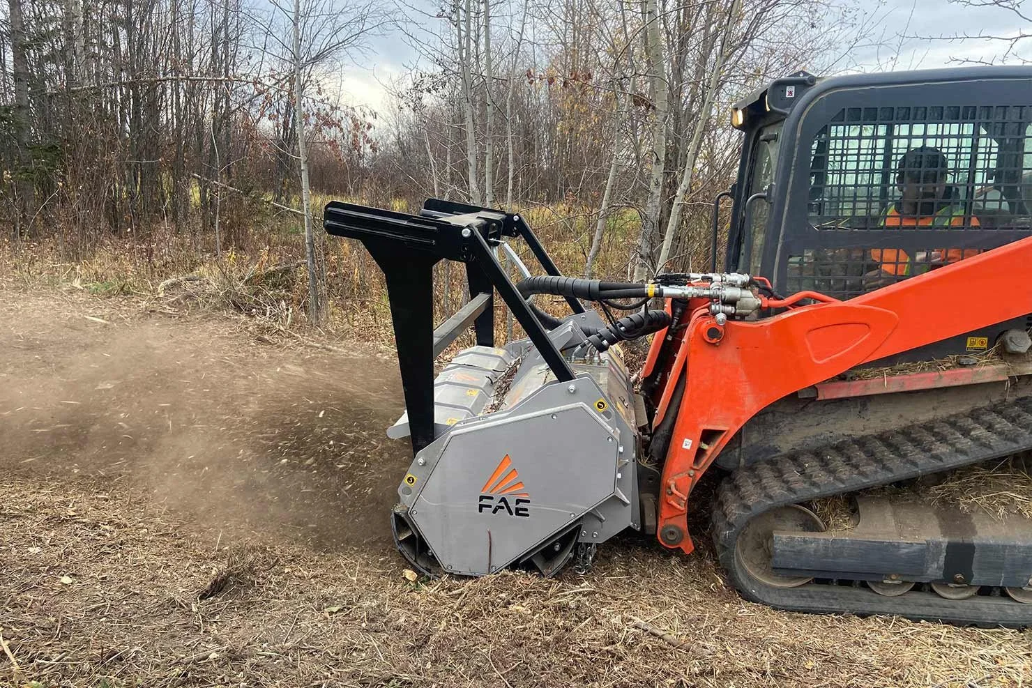 A compact excavator with an orange arm and a gray FAE mulcher head attachment working on a dirt path in a wooded area.