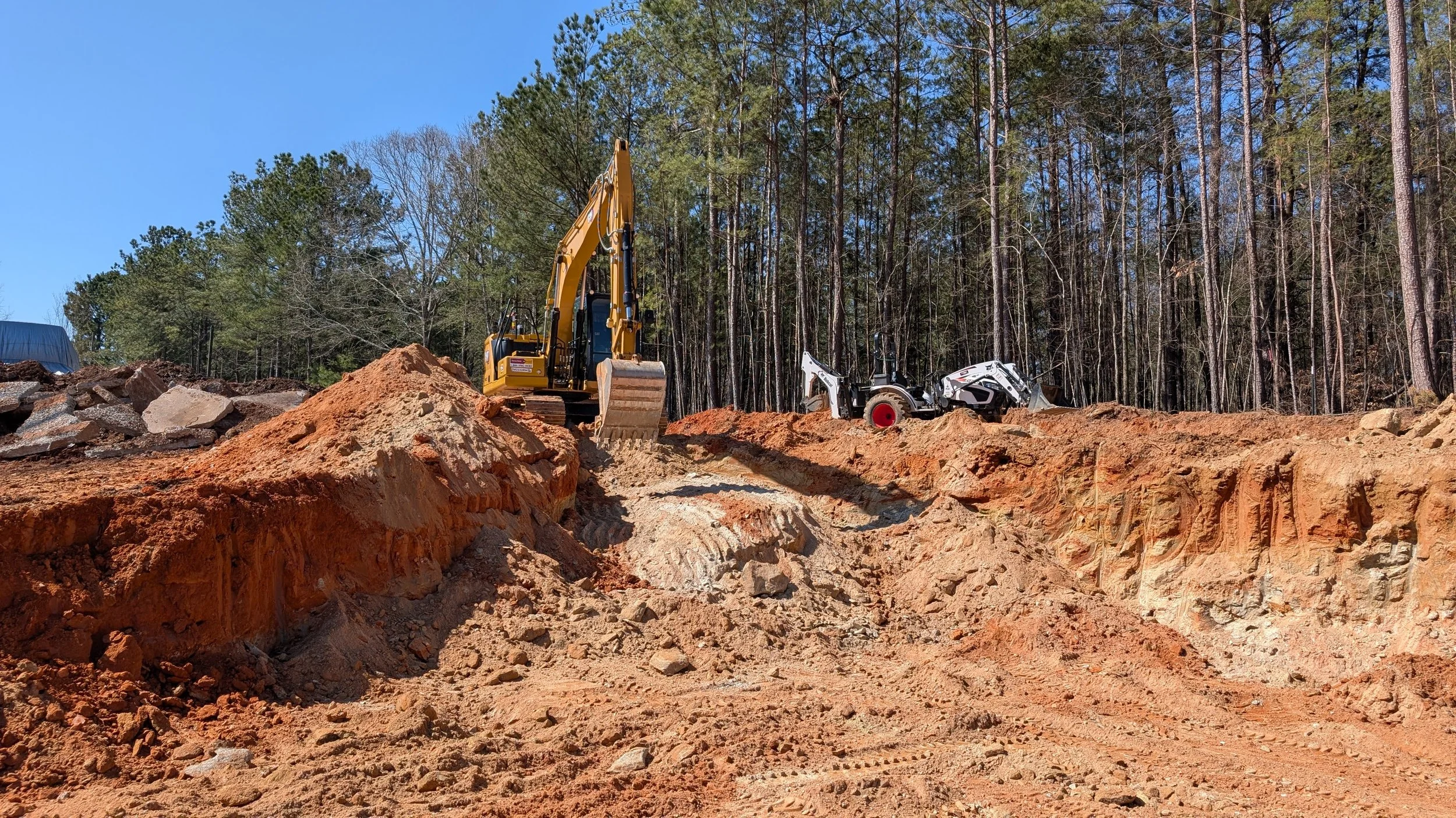 Construction site with a yellow excavator and a white compact tractor working on a dirt hill, with a forest in the background under a clear blue sky.