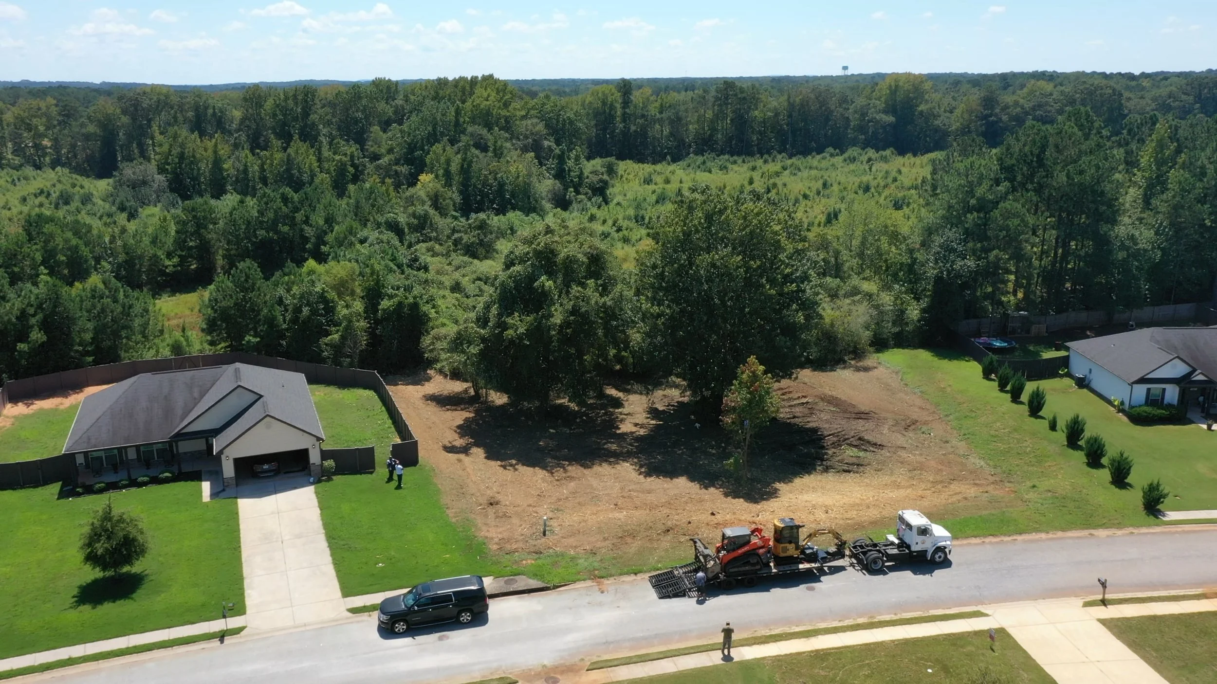 Aerial view of a residential neighborhood with two houses, a large open backyard, trees, and a construction truck on the street.