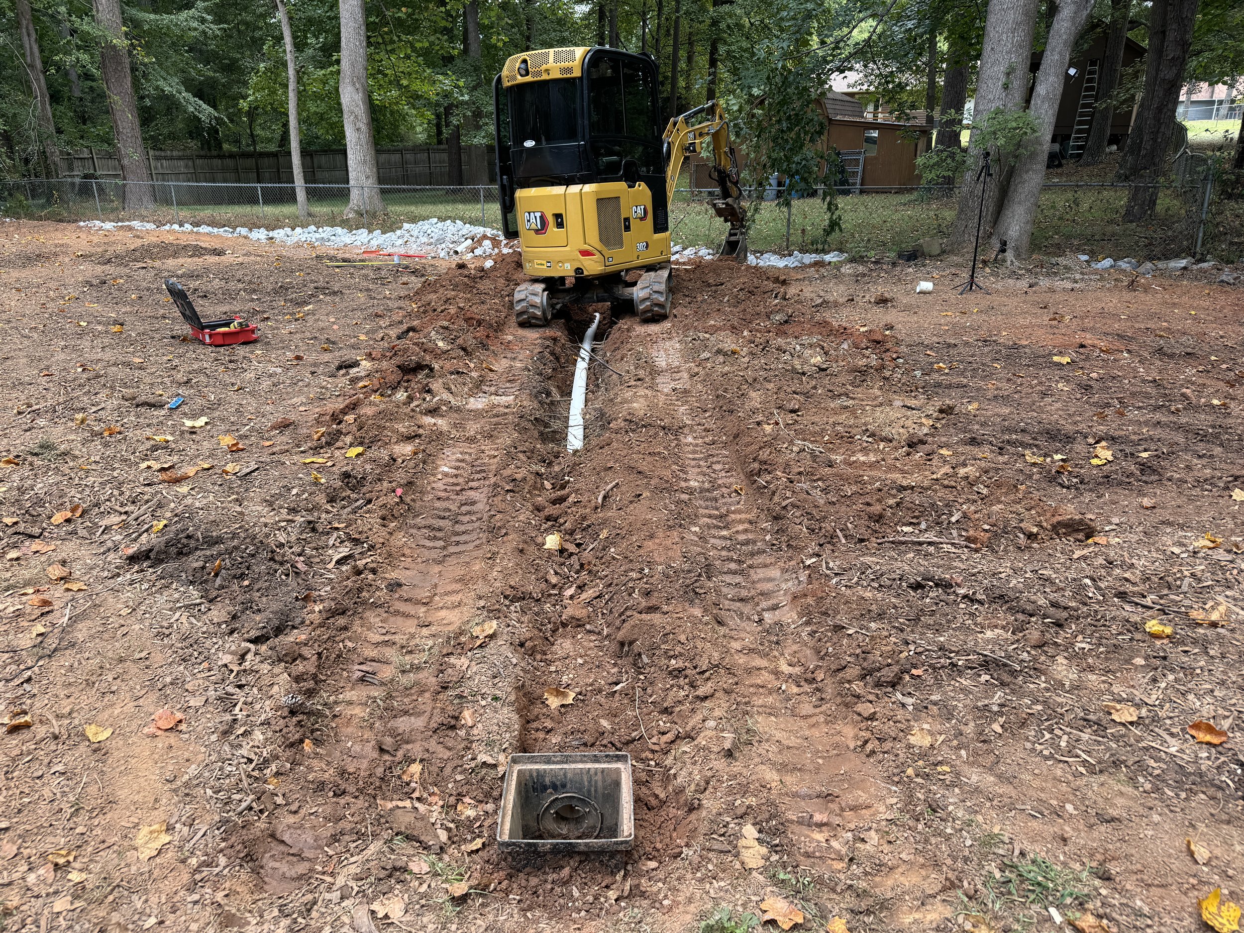 A small excavator digging a trench in a backyard, with a white pipe laid inside it. The area is muddy with tire tracks, and there is a toolbox and construction tools nearby. Trees and a wooden fence are in the background.
