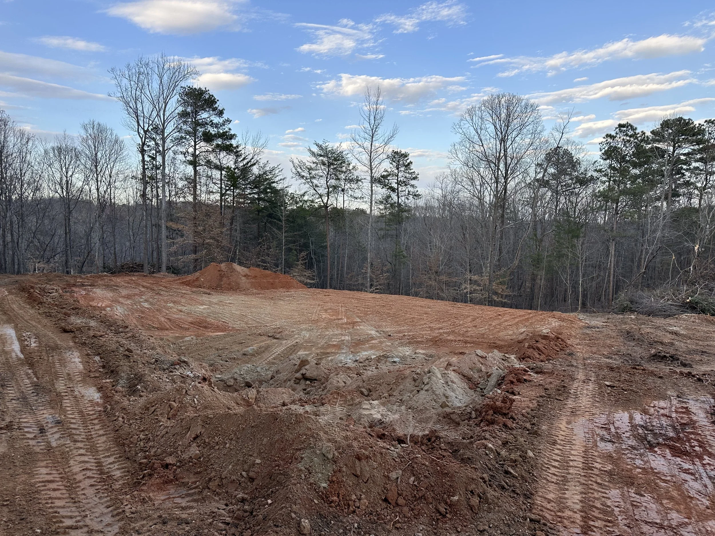 Construction site with cleared dirt and tire tracks, surrounded by leafless trees under a blue sky with scattered clouds.