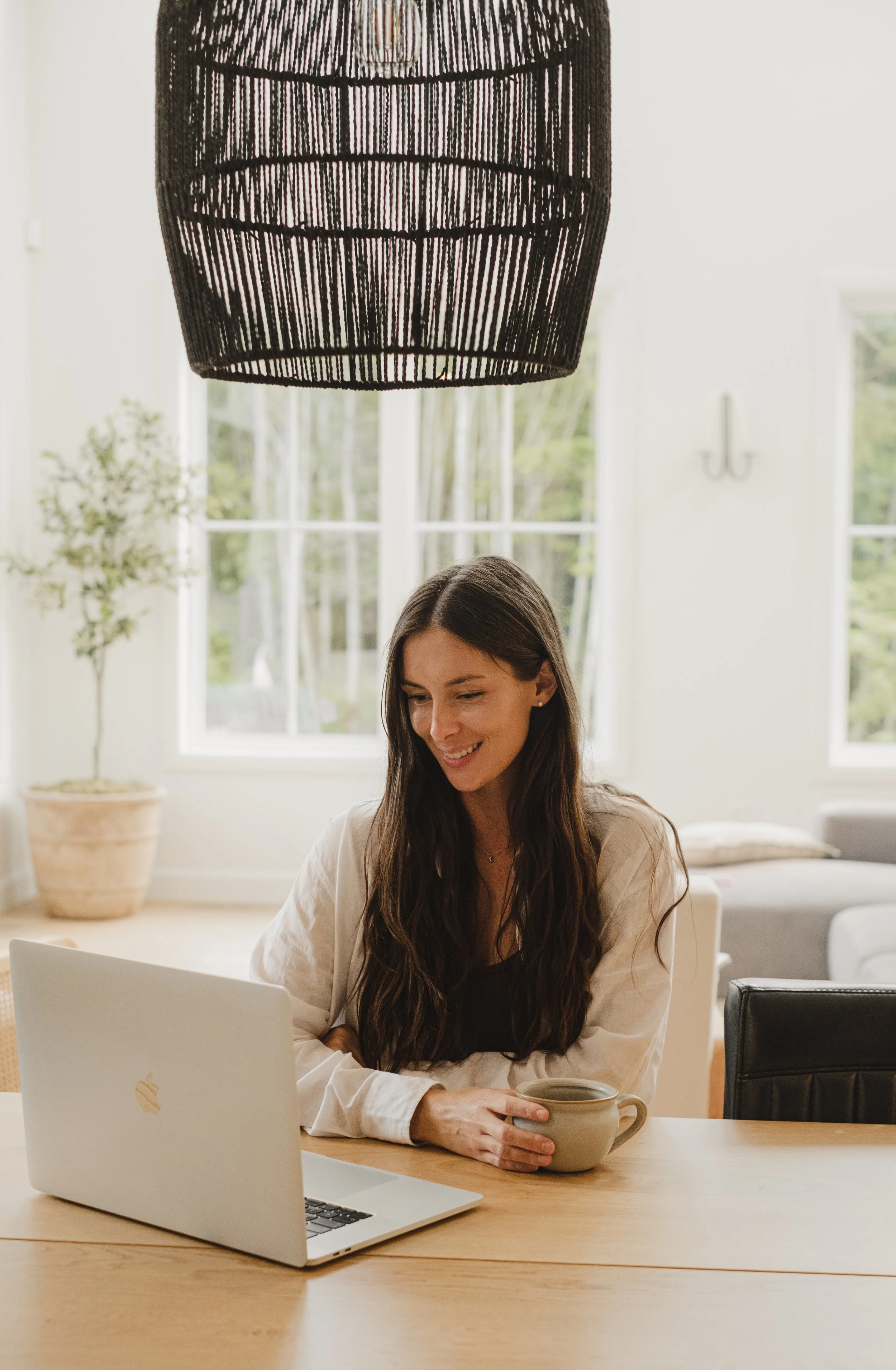 Une femme avec des cheveux longs et bruns travaille sur un ordinateur portable en souriant, assise à une table en bois dans une pièce lumineuse avec de grandes fenêtres, tenant une tasse ou un mug dans sa main, avec un intérieur moderne et des plantes en décoration.
