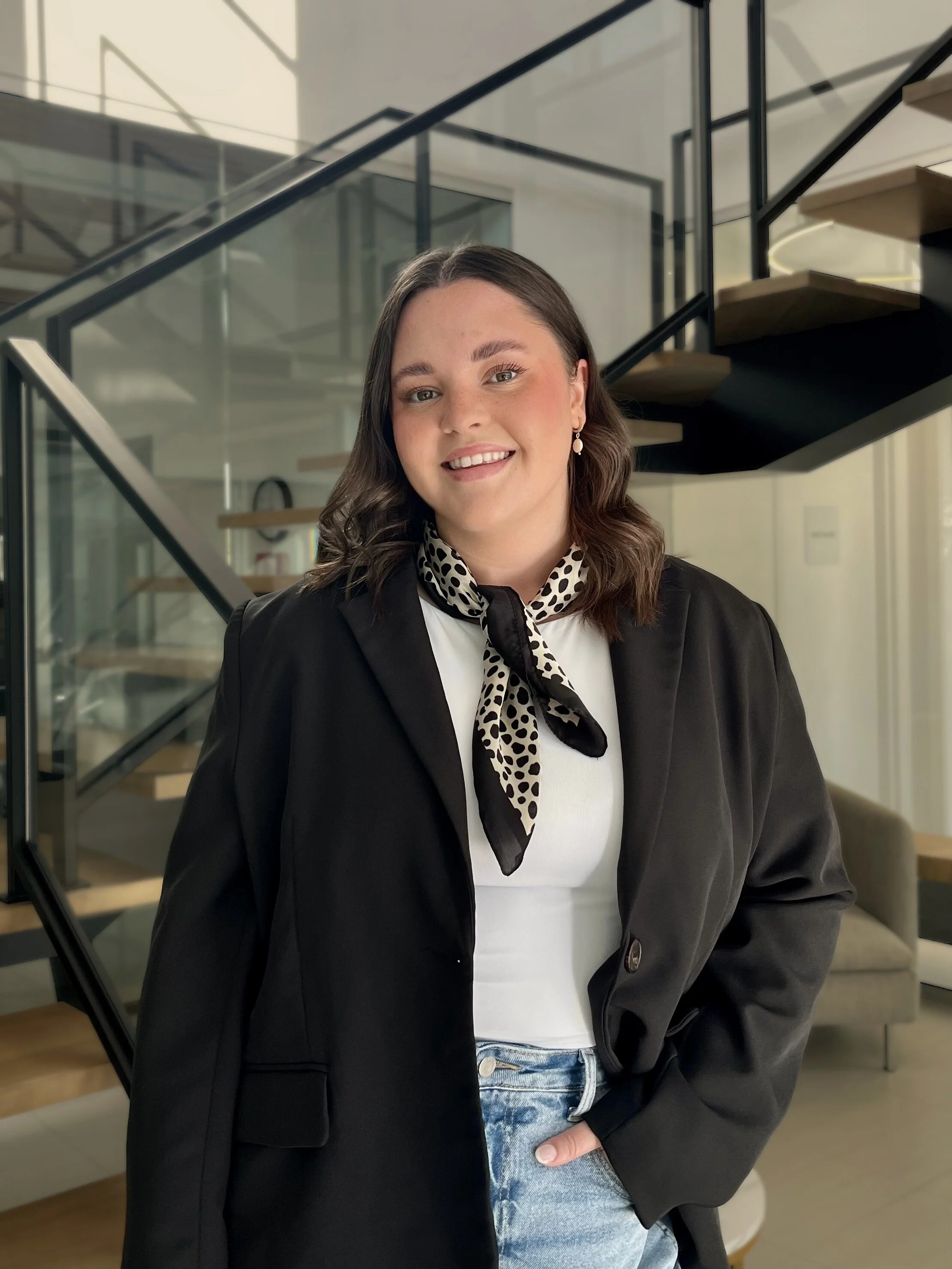 Une jeune femme souriante avec des cheveux bruns ondulés, portant une écharpe noire à pois beige, une veste noire, un t-shirt blanc, et un jean, debout dans un intérieur moderne avec un escalier en acier et bois en arrière-plan.