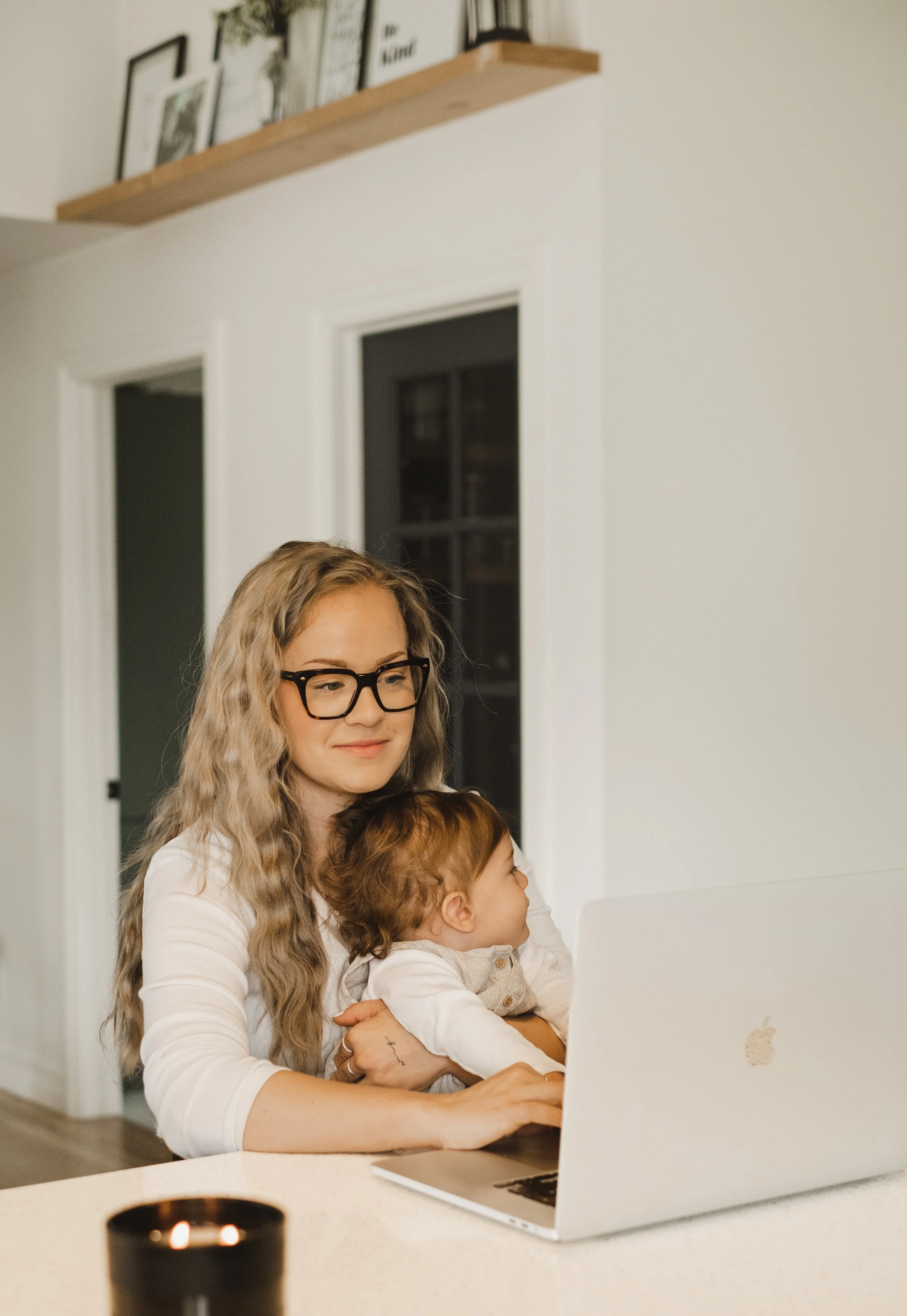 Une femme avec des lunettes et un enfant regardent un ordinateur portable blanc en utilisant une tablette, dans une cuisine moderne.