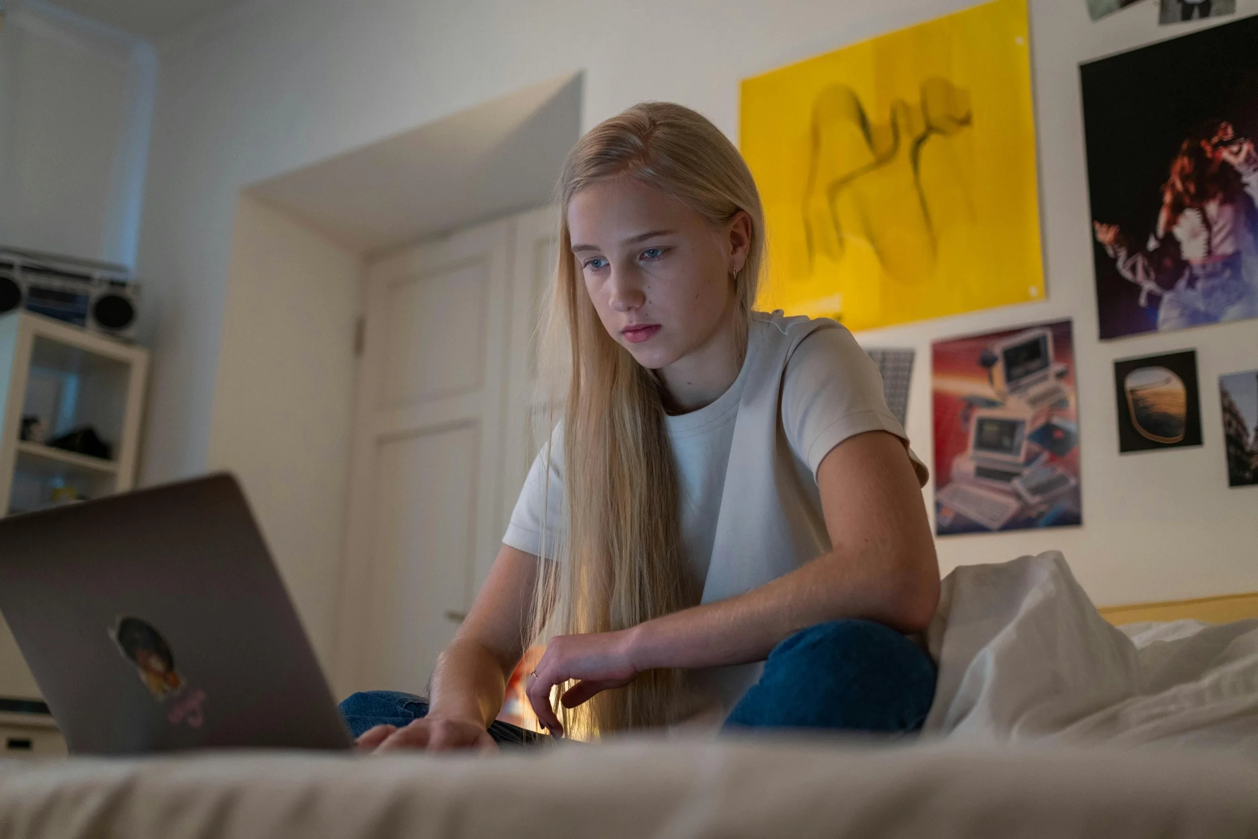 Jeune fille blonde utilisant un ordinateur portable dans sa chambre, décorée avec des posters et des œuvres d'art modernes.