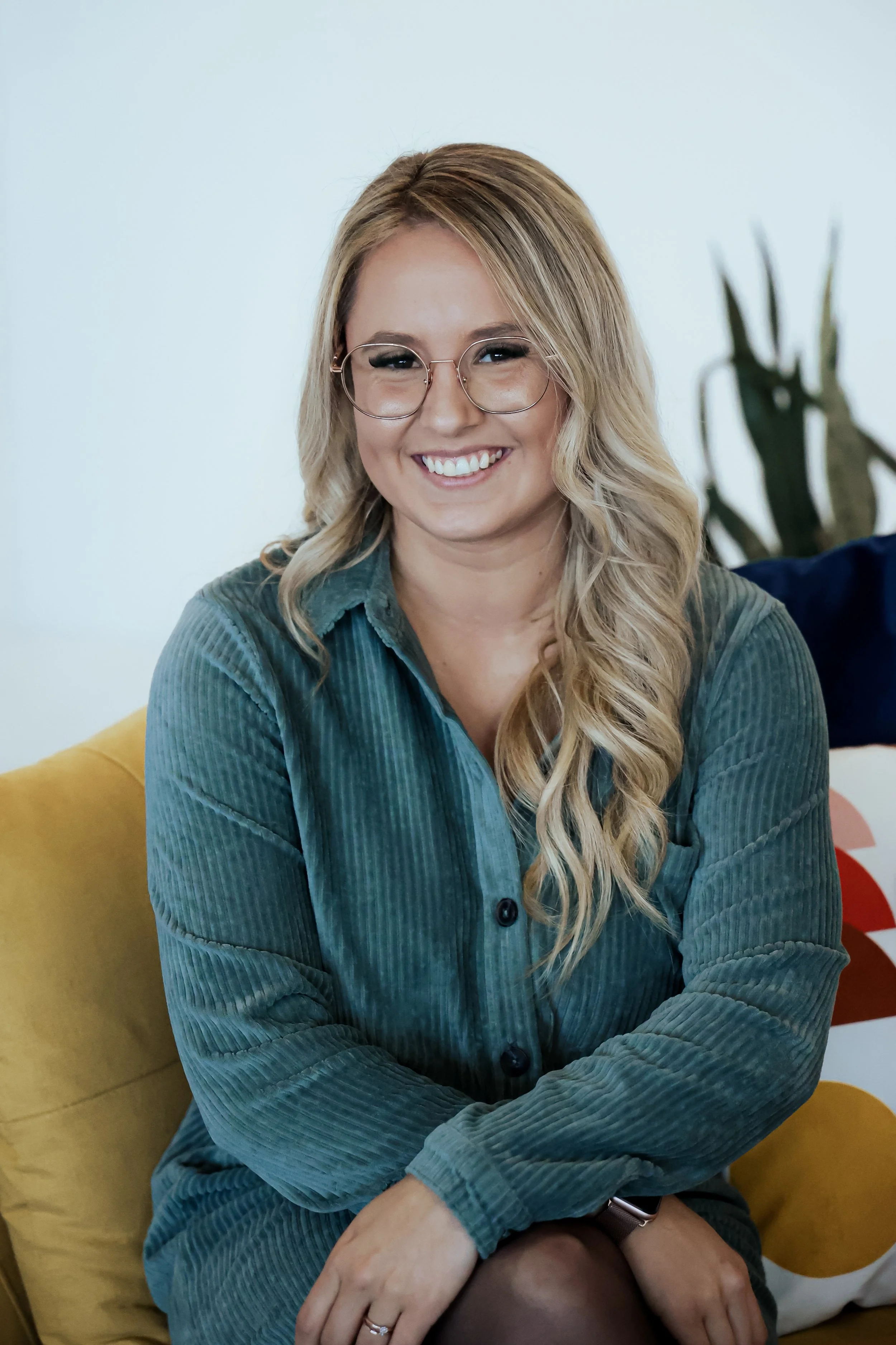 Femme souriante portant des lunettes, assise sur un canapé jaune, fond blanc avec une plante verte en arrière-plan.