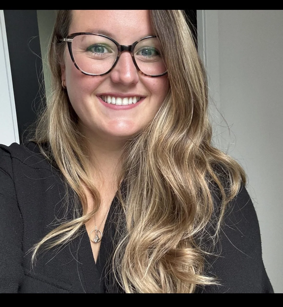 Jeune femme souriante avec des lunettes rondes, cheveux longs ondulés, porte un collier en forme de cercle, habillée en noir, pose à l'intérieur.