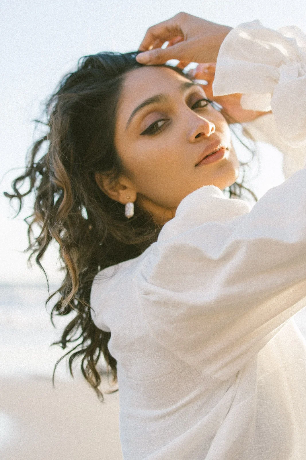 Jeune femme avec des cheveux bouclés, portant des boucles d'oreilles en argent et un chemisier blanc, posant sous la lumière naturelle.