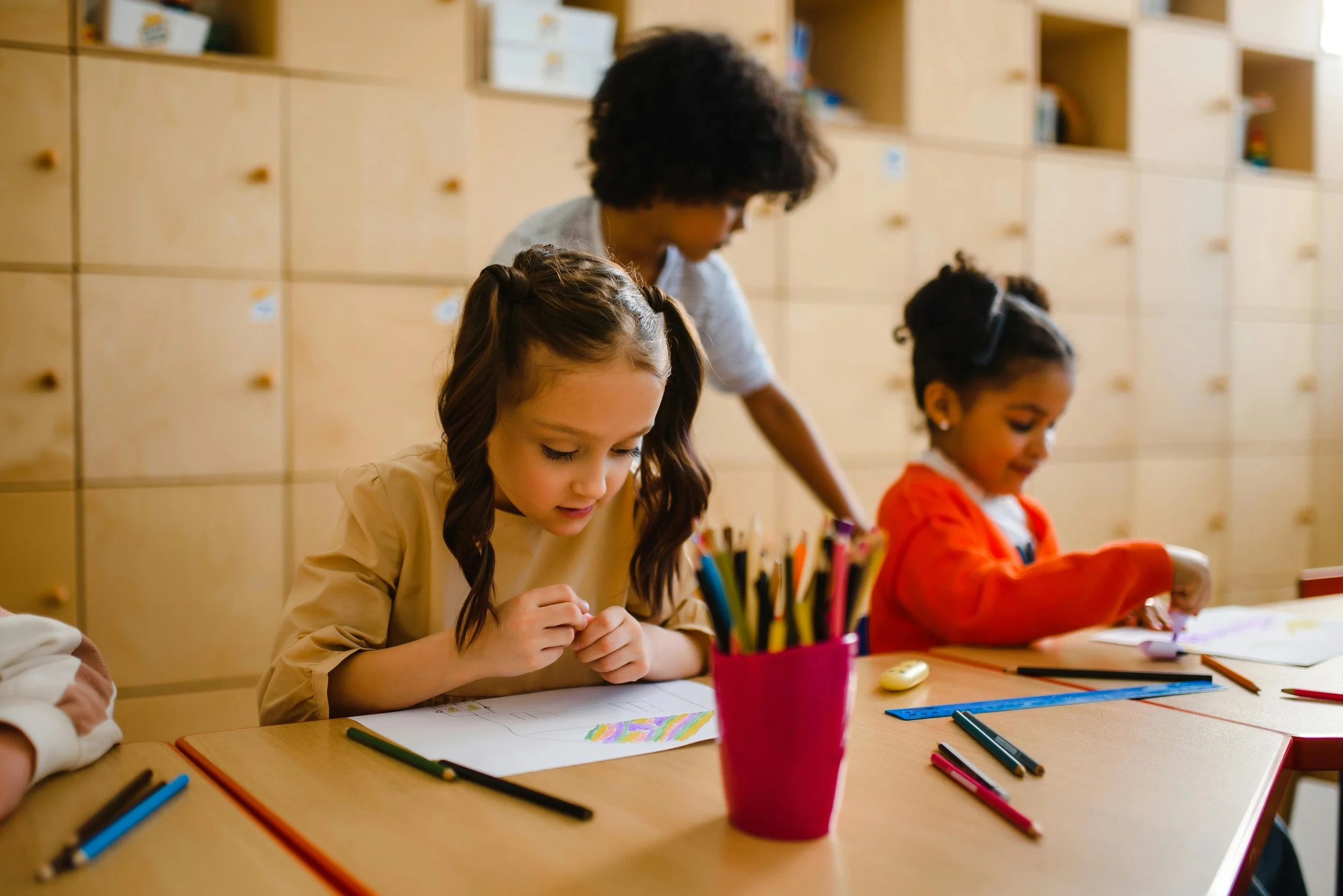 Trois enfants en classe, dessinant à leur table, avec un adulte supervisant, crayons et matériel artistique sur la table.