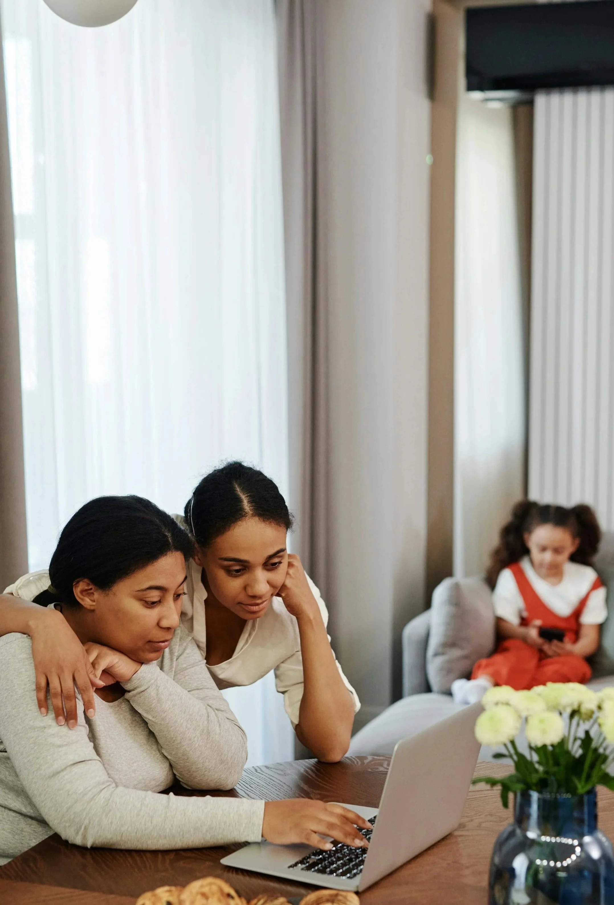 Deux femmes regardant un ordinateur portable sur une table, avec une fille assise en arrière-plan sur un canapé.