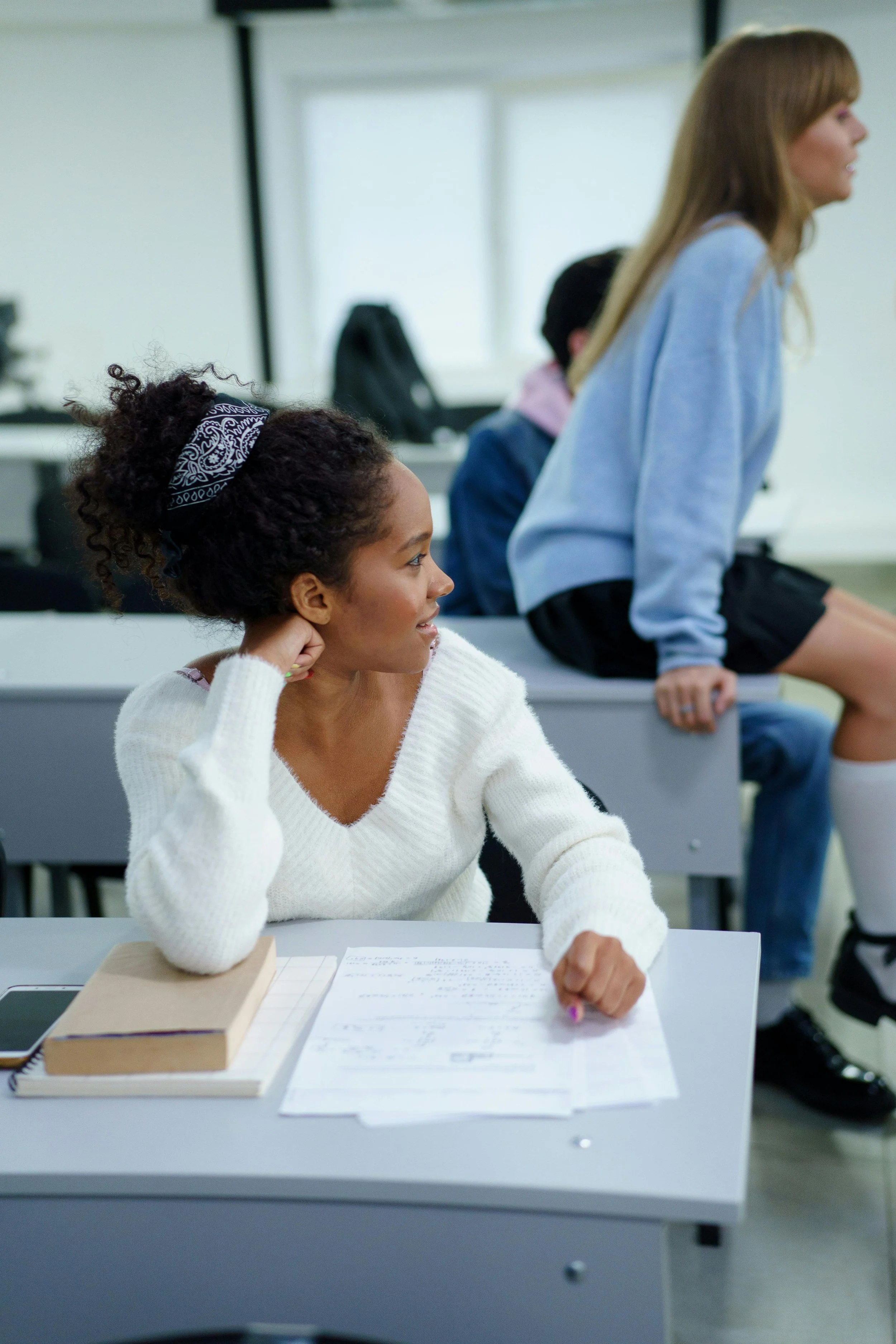 Une étudiante africaine à la chevelure bouclée et portant un pull blanc, assise à un bureau avec des notes et un téléphone portable devant elle, dans une salle de classe en train d'écouter une autre étudiante qui est debout à côté d'elle.