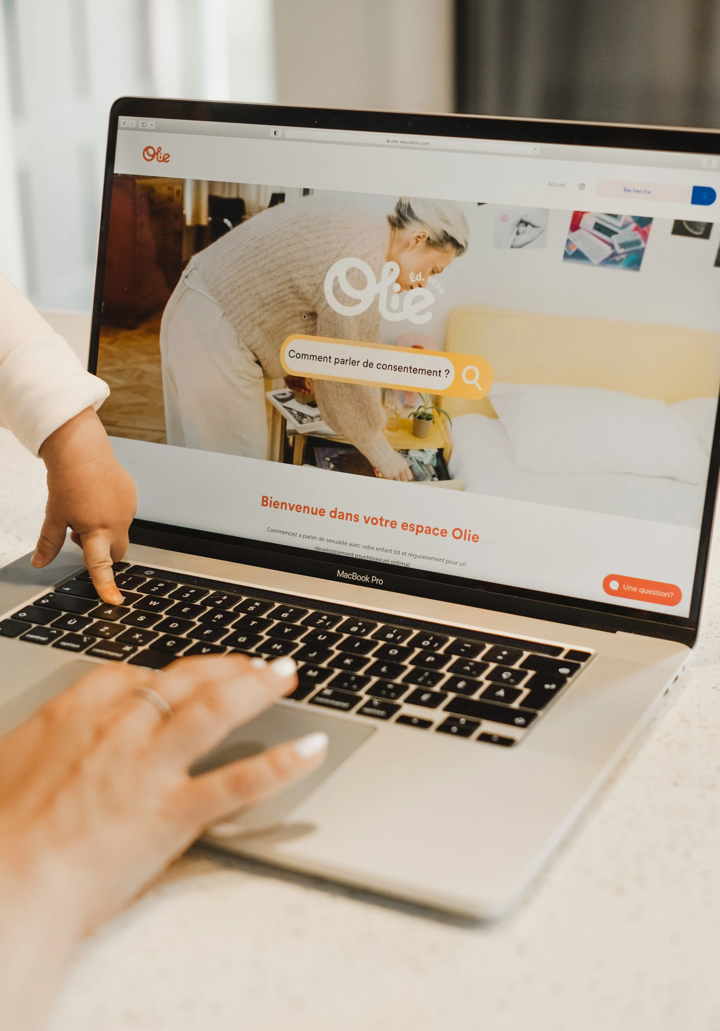 Capture d'écran d'un site web nommé 'Olie' avec une barre de recherche demandant 'Comment parler de consentement ?', affiché sur un ordinateur portable MacBook Pro. Une personne pointe avec son doigt sur le clavier de l'ordinateur.