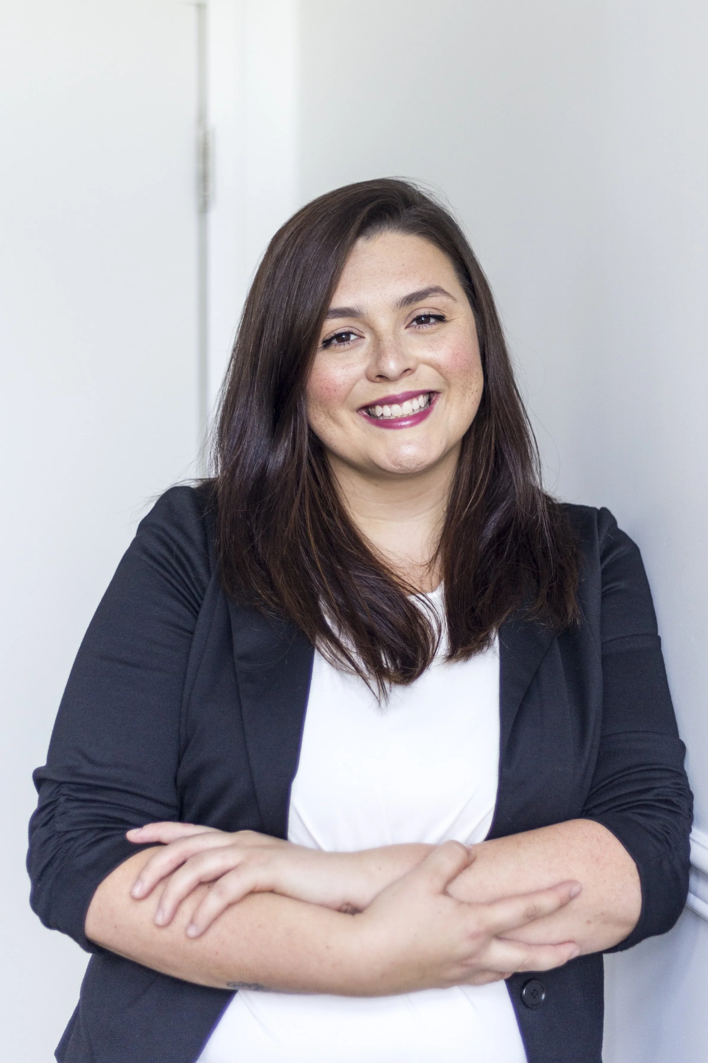 Jeune femme souriante avec des bras croisés, portant une veste noire et un haut blanc, debout contre un mur blanc.
