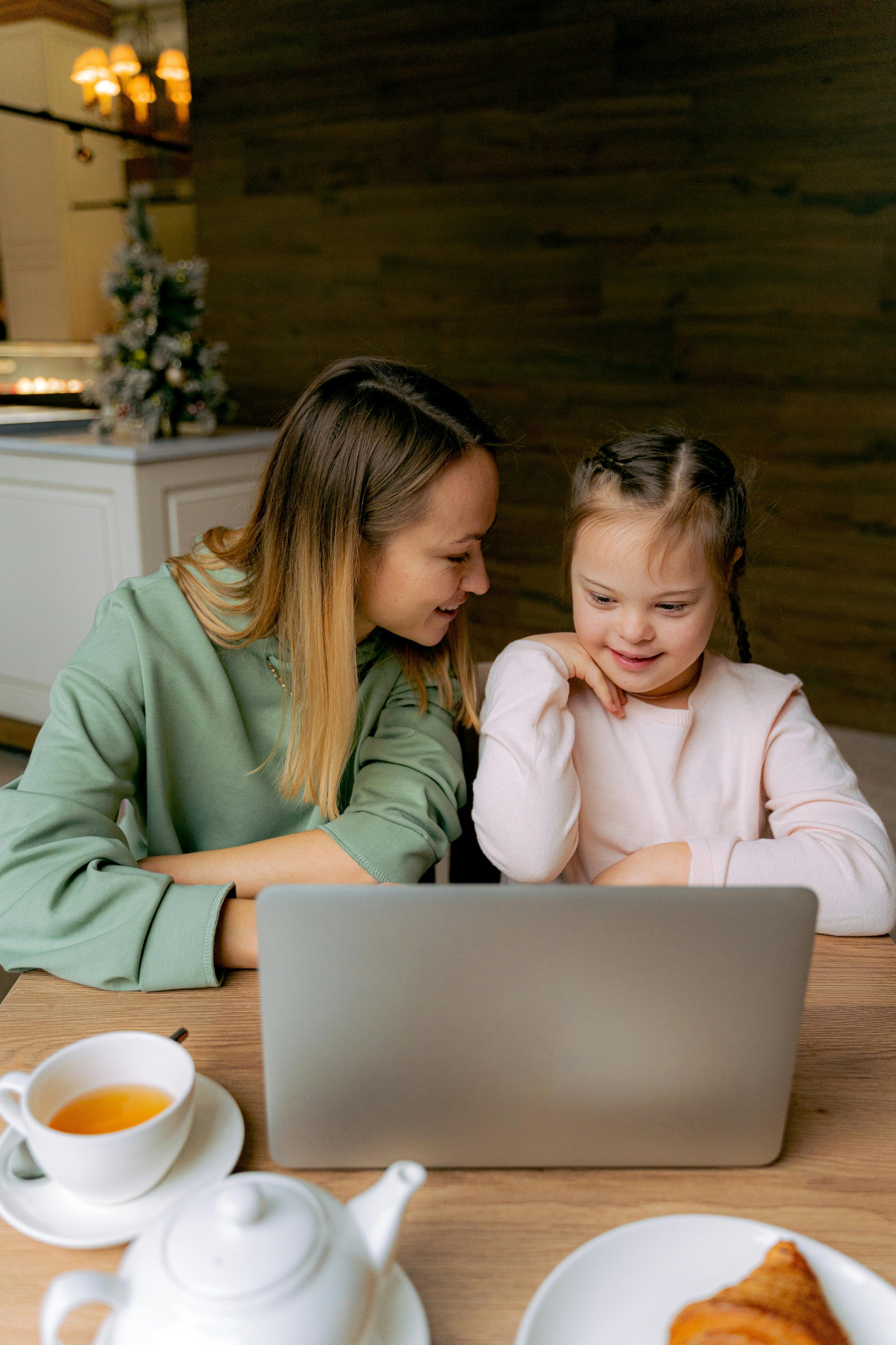 Une femme et une jeune fille regardent un ordinateur portable ensemble à une table dans une cuisine, avec une tasse de thé et un croissant sur la table.