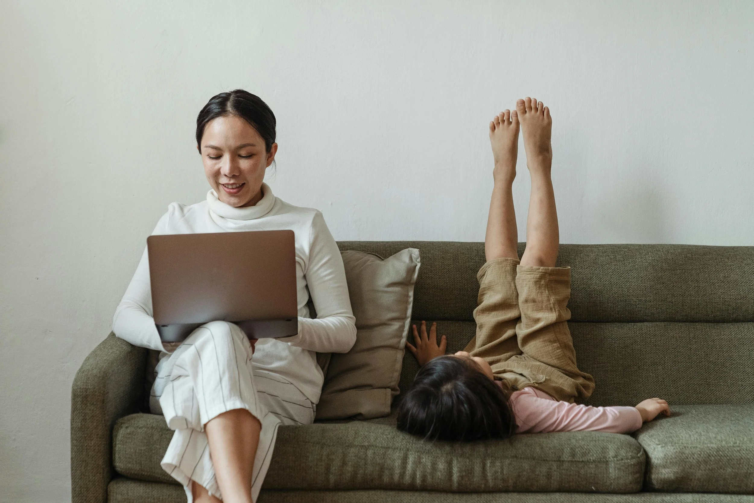 Une femme assise sur un canapé avec un ordinateur portable sur ses genoux, regardant l'écran et souriant; un enfant allongé sur le dos à côté d'elle avec les jambes levées contre le mur, dans une pièce au mur blanc.