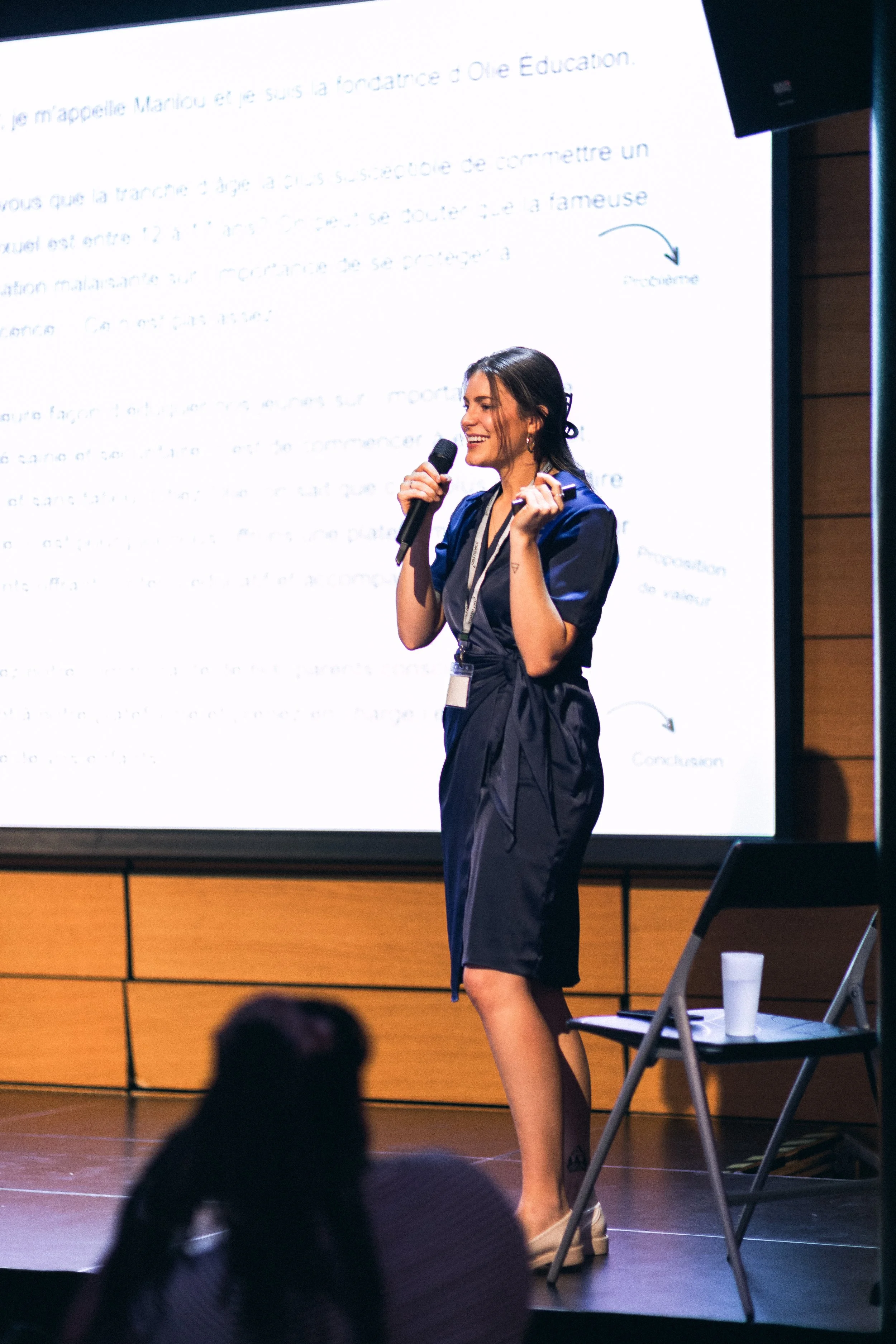 Femme souriante en robe bleue parlant lors d'une présentation avec un micro, dans une salle de conférence, projection en arrière-plan.