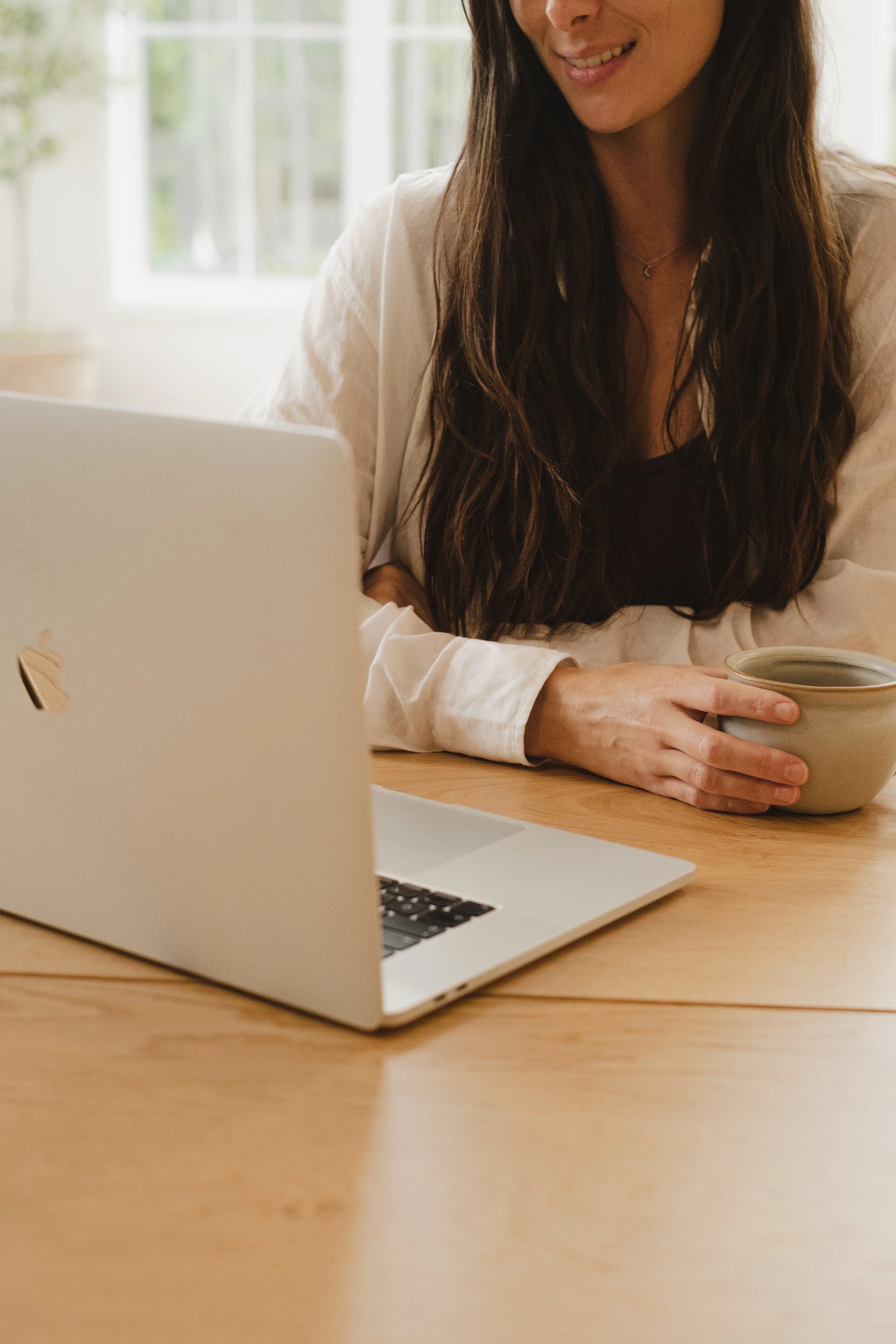 Femme assise à un bureau en train de regarder un ordinateur portable, tenant un bol et souriant, dans une pièce lumineuse.