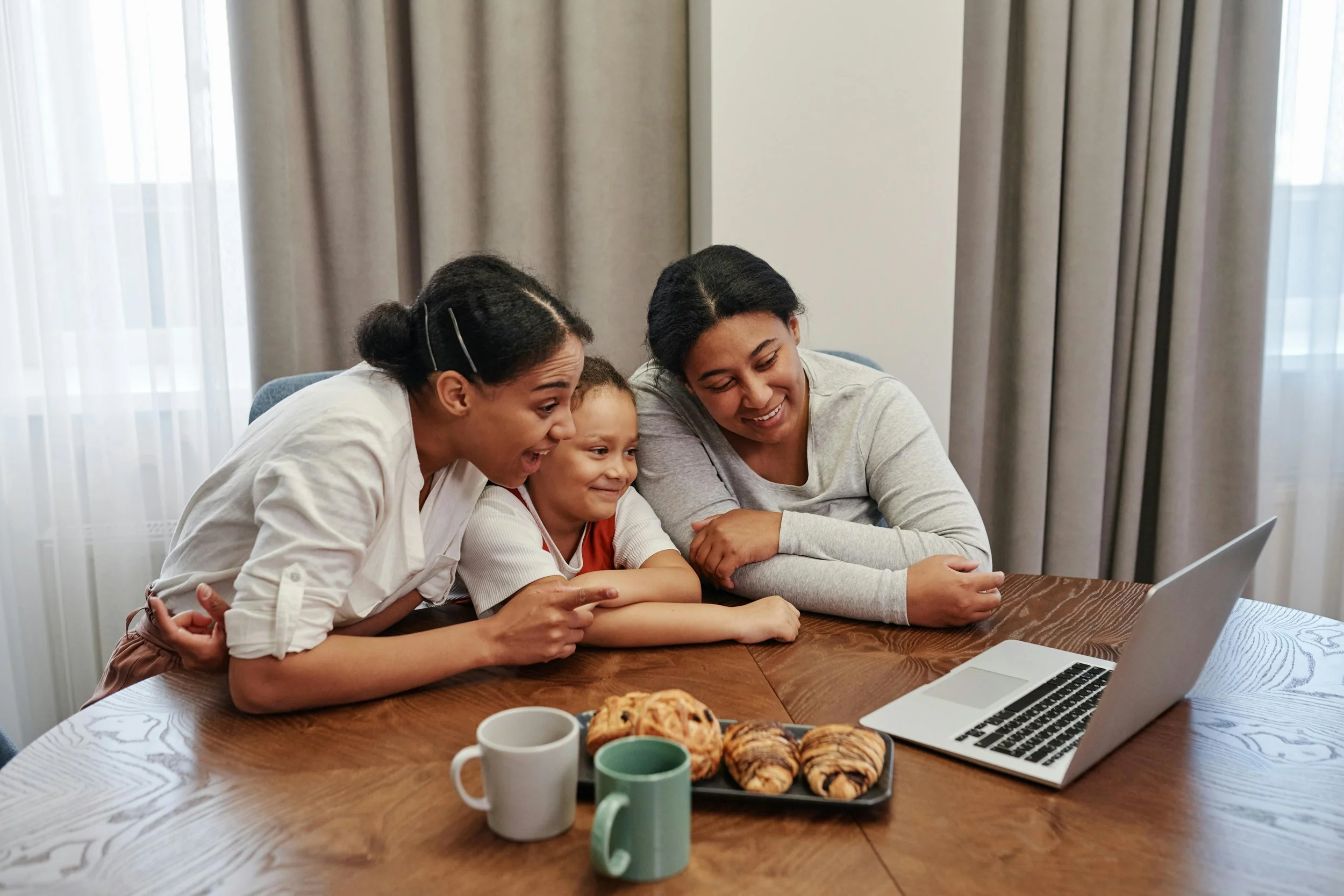 Trois femmes et un enfant regardent avec enthousiasme un ordinateur portable sur une table en bois, avec des croissants, deux mugs, et un bol à côté, dans une pièce éclairée par la lumière naturelle.