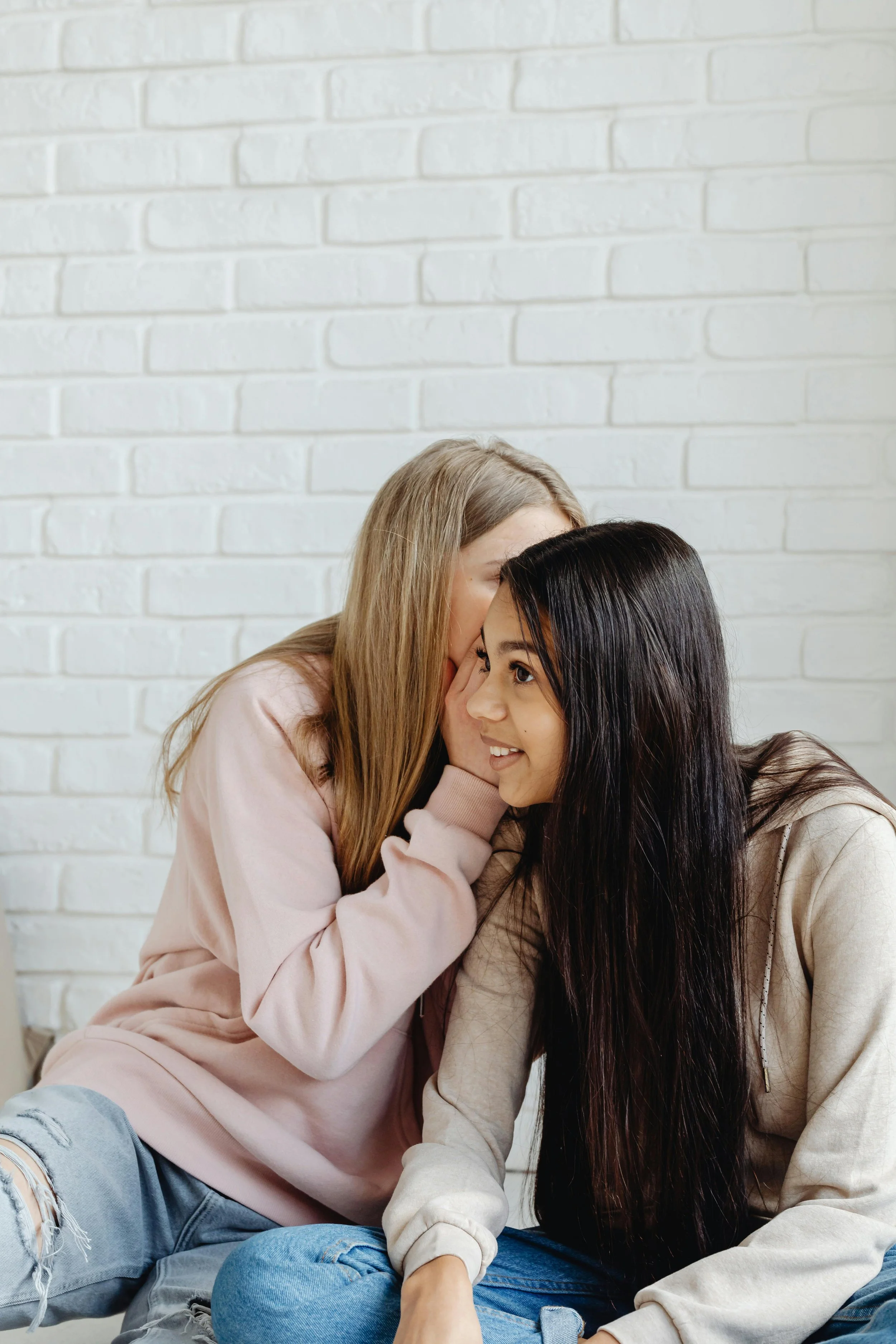 Deux jeunes filles assises près d'un mur blanc, souriantes et partageant un moment de complicité.