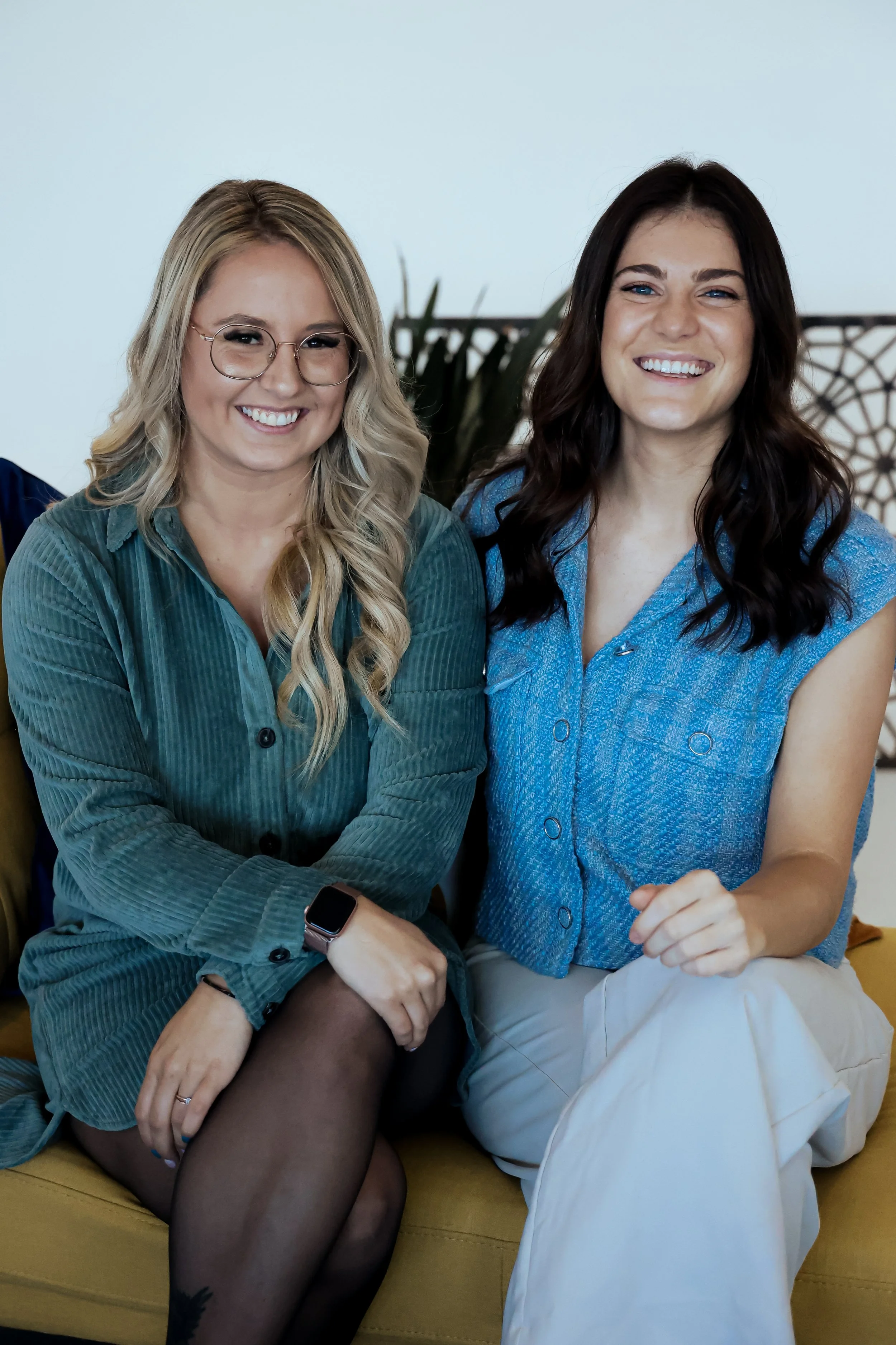 Deux femmes souriantes assises côte à côte sur un canapé, l'une porte des lunettes et une veste vert foncé, l'autre porte un haut bleu et des pantalons clairs.