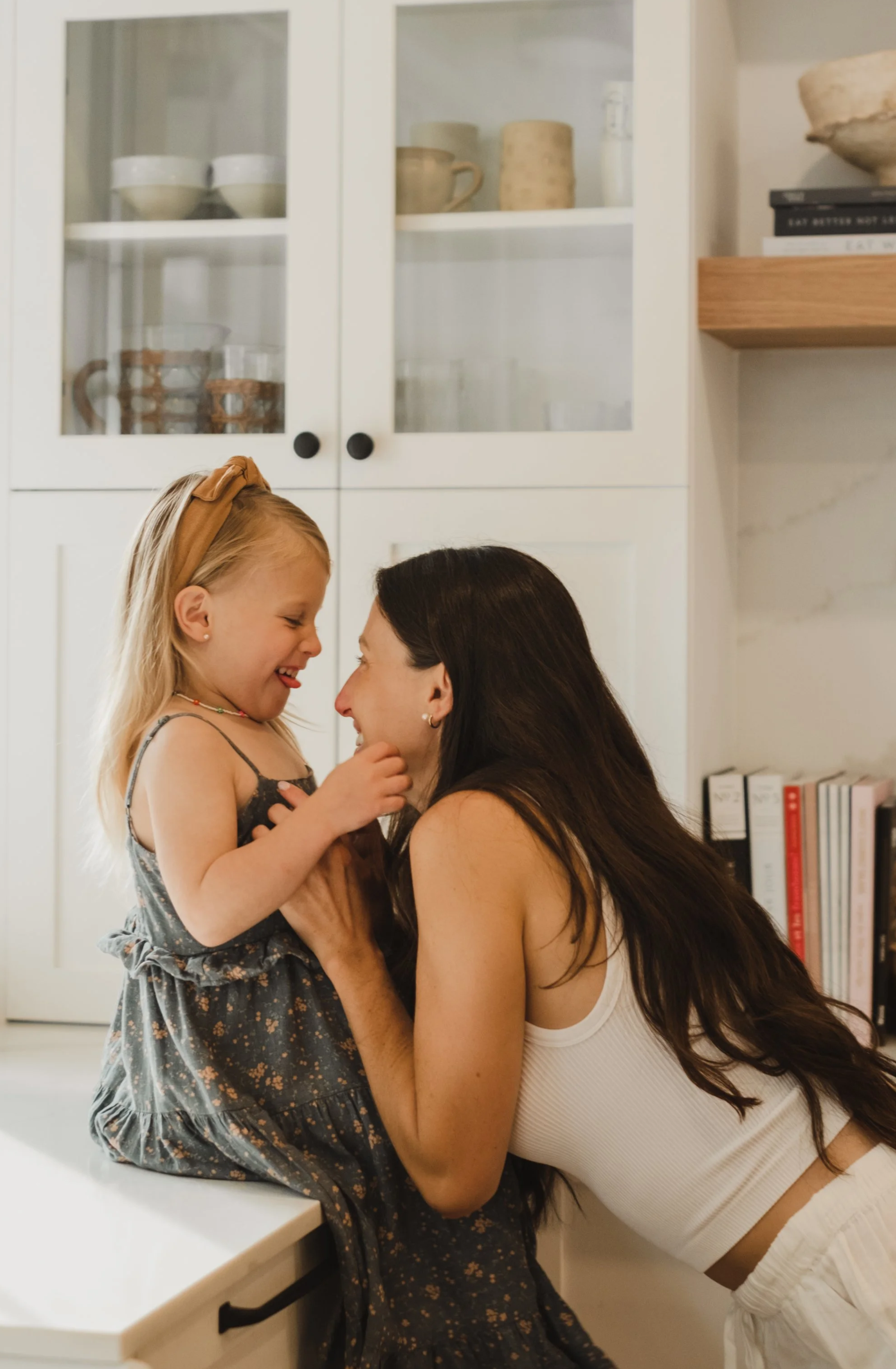 Une mère et sa fille rient et jouent ensemble dans une cuisine, face à face, avec des étagères de vaisselle en arrière-plan.