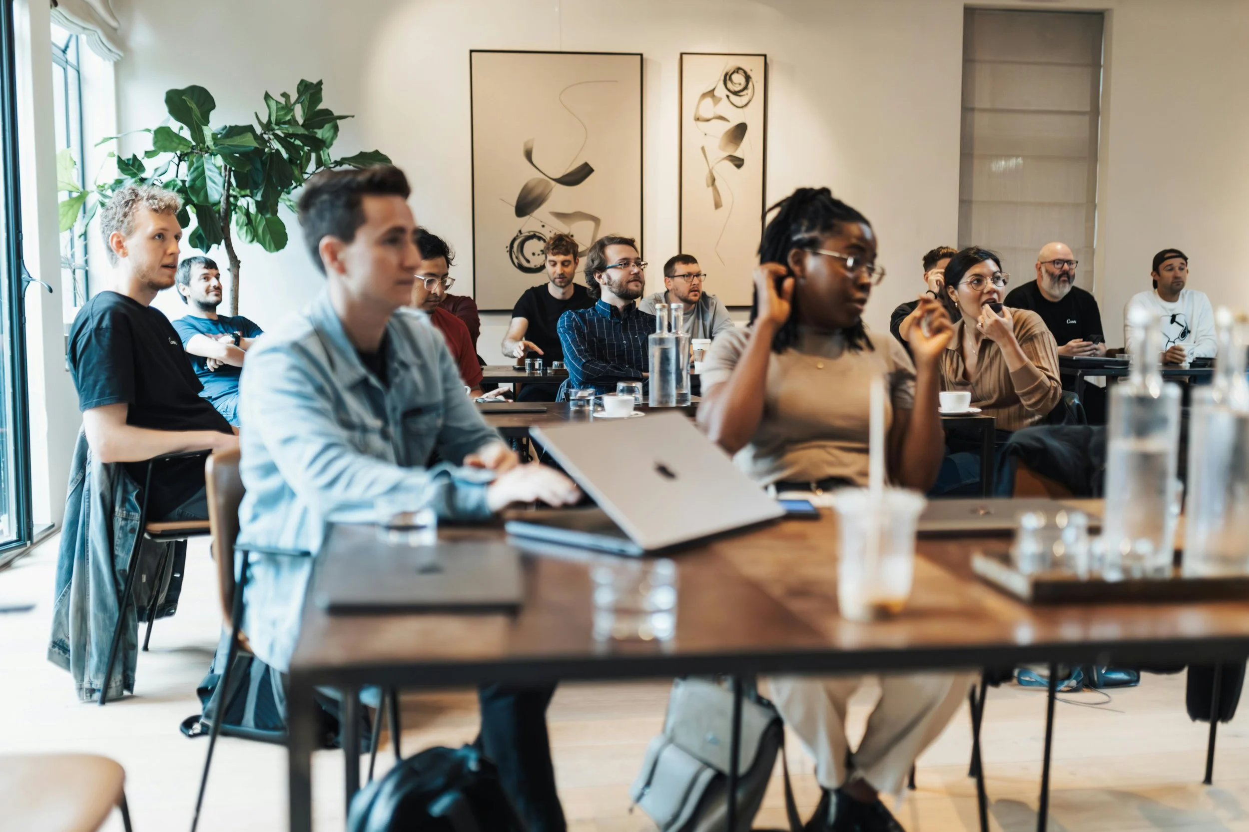 Groupe de personnes assises dans une salle de réunion ou un atelier discussions variées, certains prennent des notes ou regardent vers l'avant.