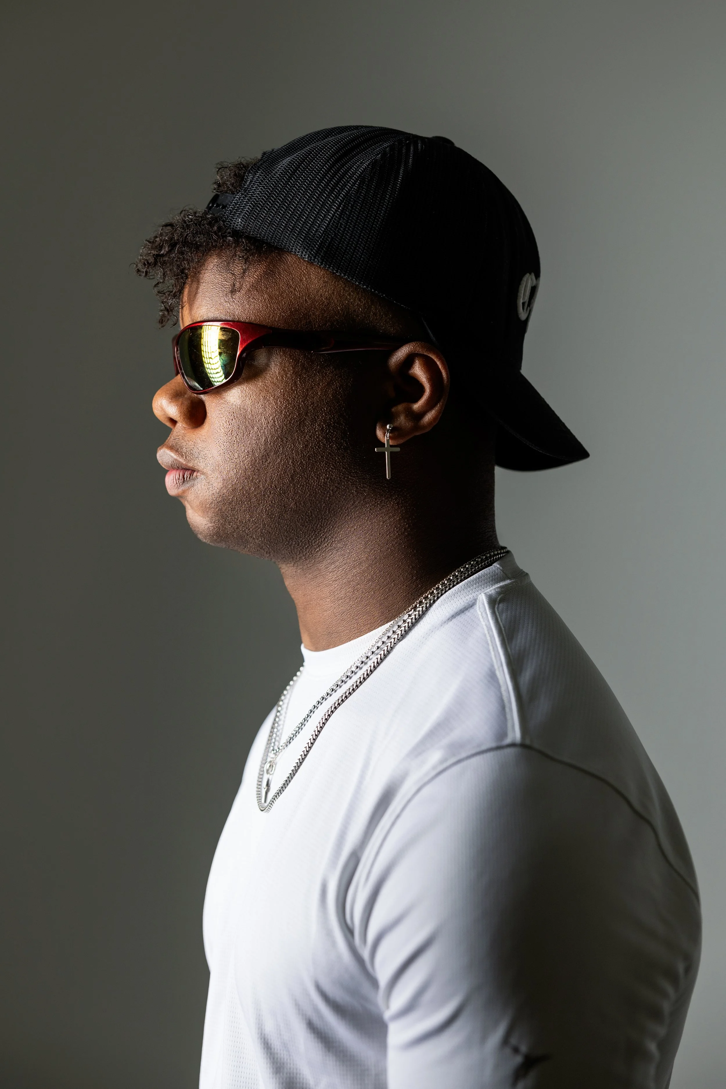 Profile of a young man wearing a black cap backwards, red sunglasses, cross earring, silver chains, and a white athletic shirt, against a gray background.
