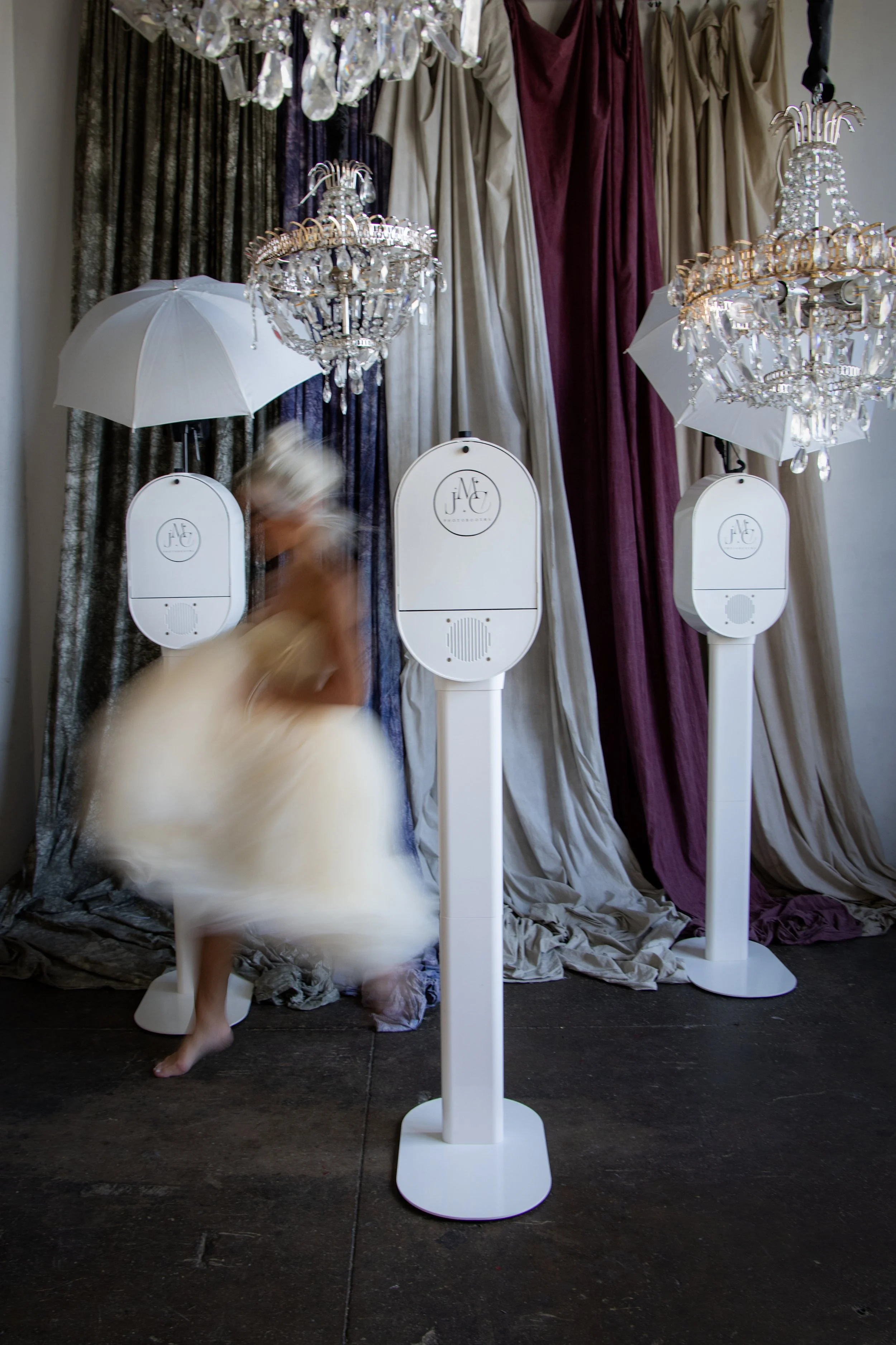A blurred woman in a white dress appears to be dancing or moving inside a decorative setting with chandeliers, white umbrellas, and large draped curtains.