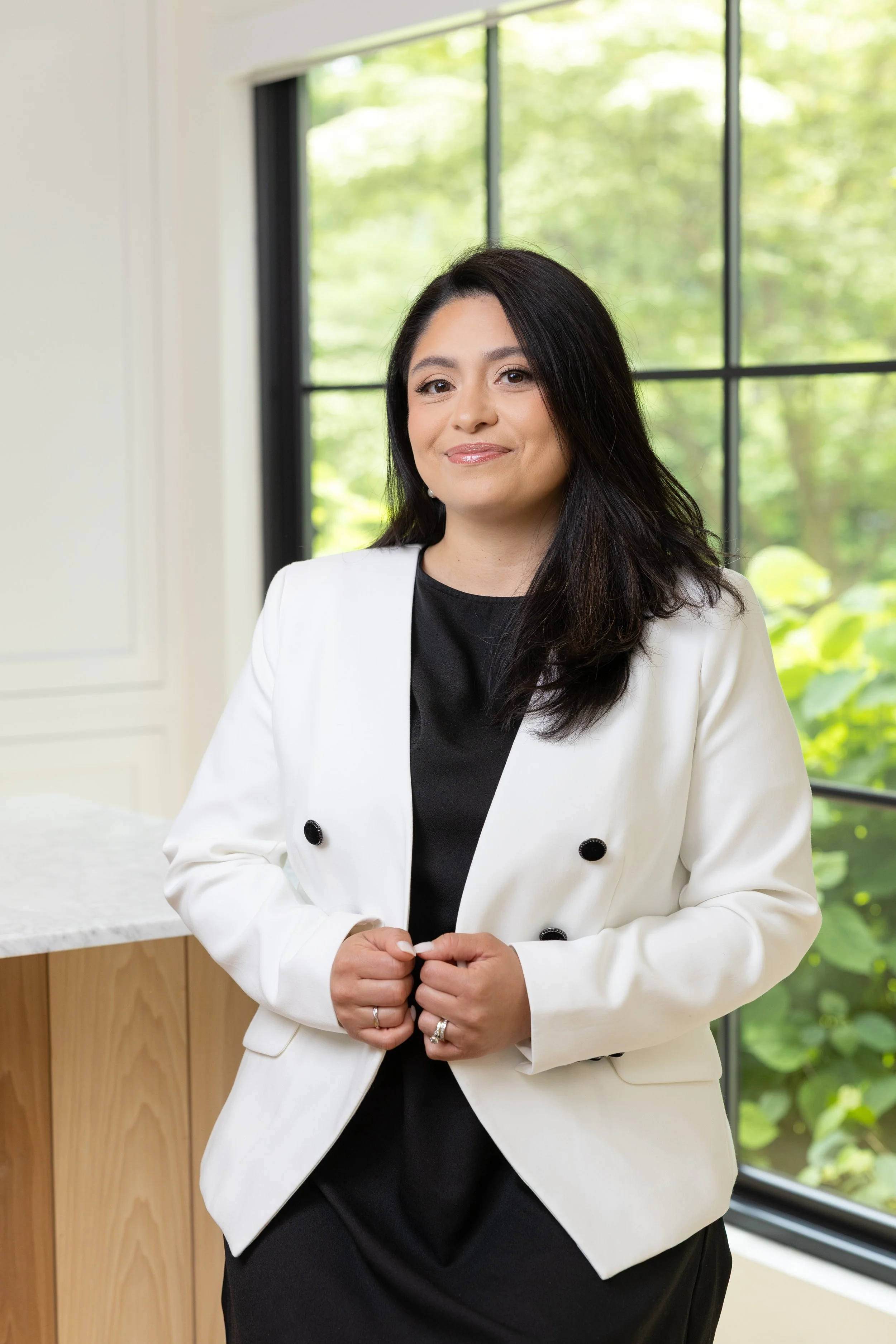 A woman with long dark hair wearing a white blazer over a black top, standing indoors in front of a large window with a green outdoor view.