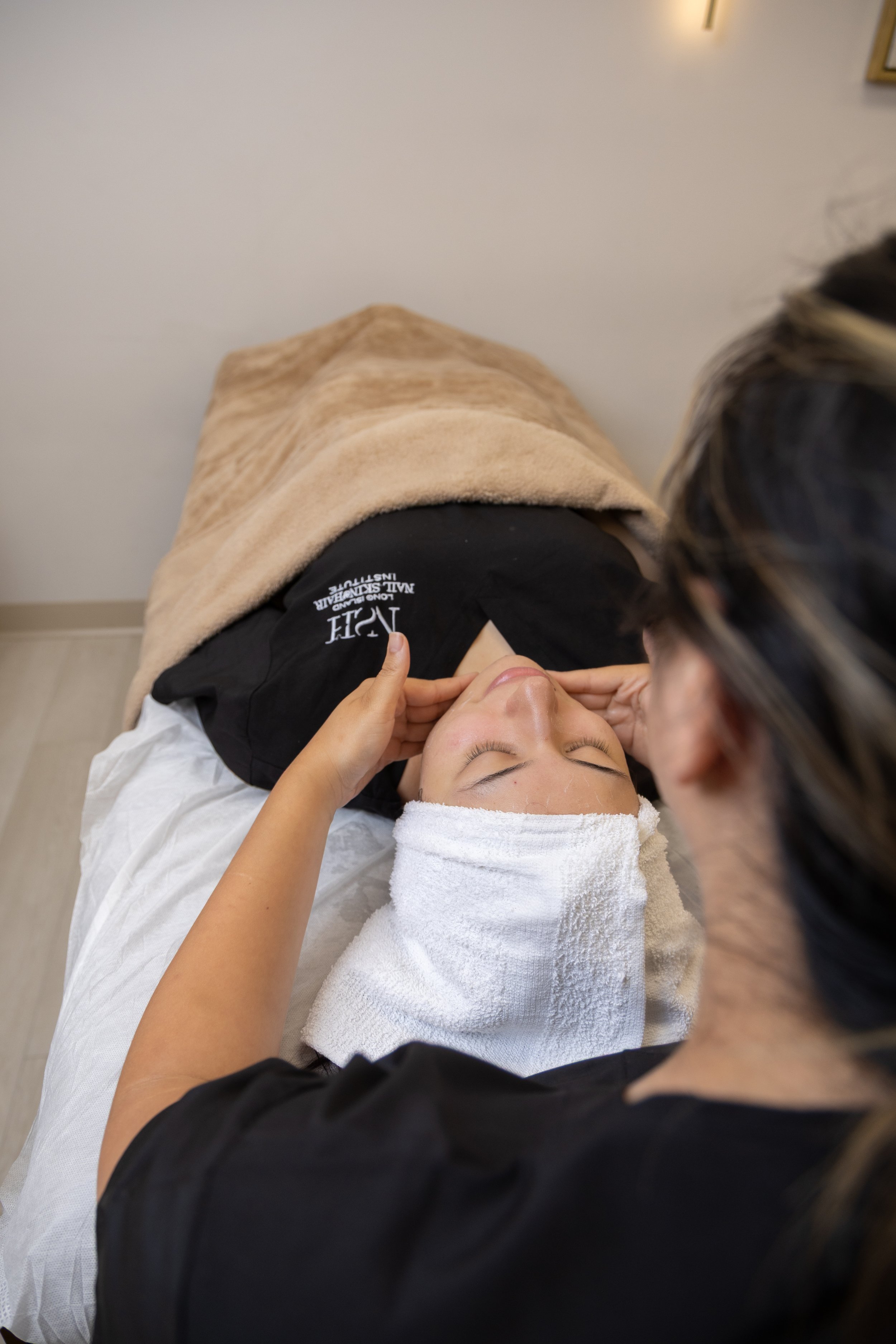 A woman receiving a facial treatment in a spa setting, lying on a massage table with a towel wrapped around her head and face, while a professional applies a facial treatment.