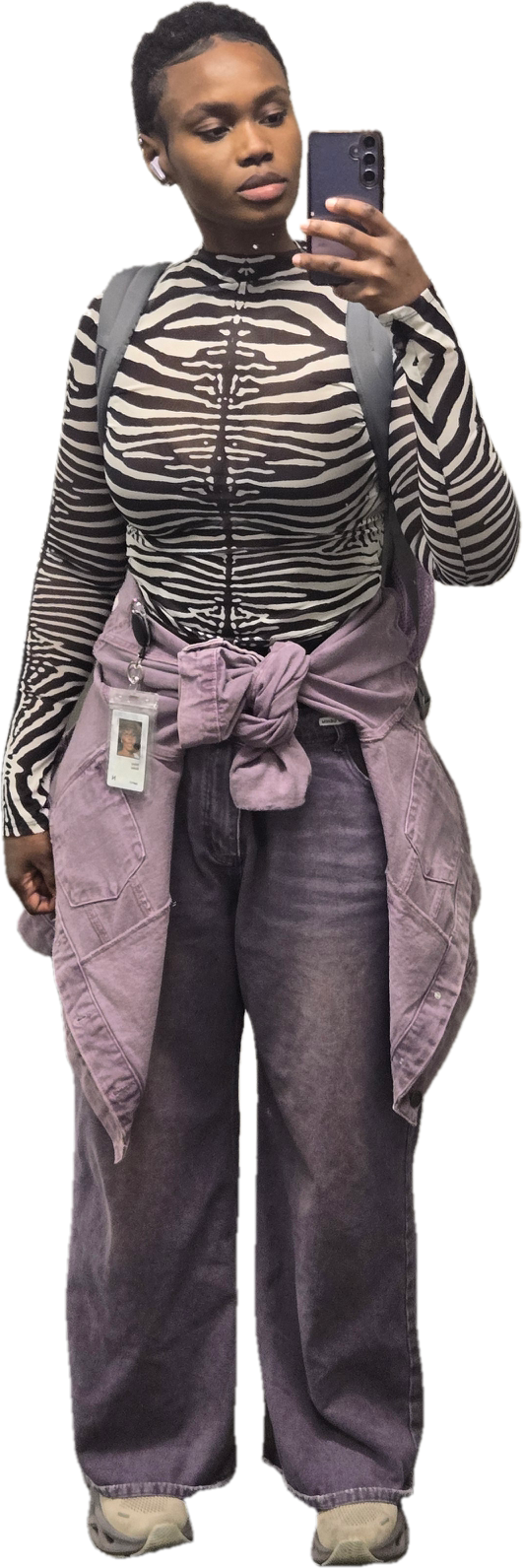 Young woman taking a mirror selfie with her phone, wearing a zebra print long-sleeve top, gray jeans, and a purple jacket tied around her waist with a backpack over one shoulder.