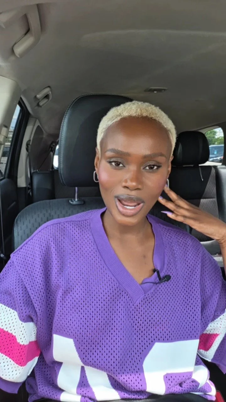 Hilary Biaku | Black Female Model, Creative Strategist woman with short platinum blonde hair wearing a purple mesh sports jersey with white and pink accents, sitting inside a vehicle, touching her ear with her left hand, with a confident expression.