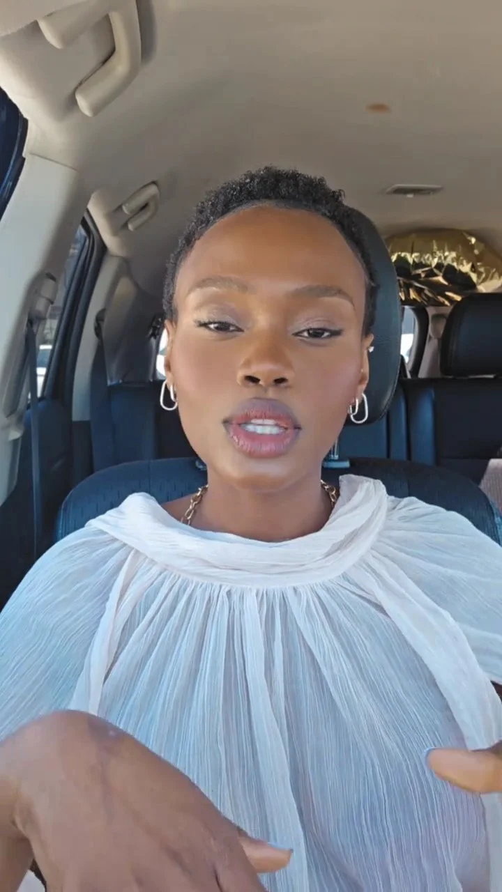 Hilary Biaku | Black Female Model, Creative Strategist, Content Creator, Public Speaker, and Life Coach — A woman sitting inside a vehicle, wearing a white pleated top, with short curly hair and hoop earrings, looking at the camera.