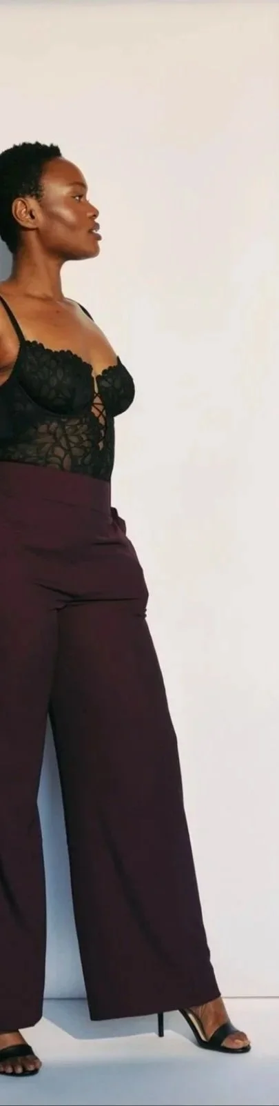 Hilary Biaku | Creative Strategist, Model, Content Creator, Public Speaker, and Life Coach — black A woman standing in profile against a plain white background, wearing a black lace top and high-waisted maroon wide-leg pants with black high heels.