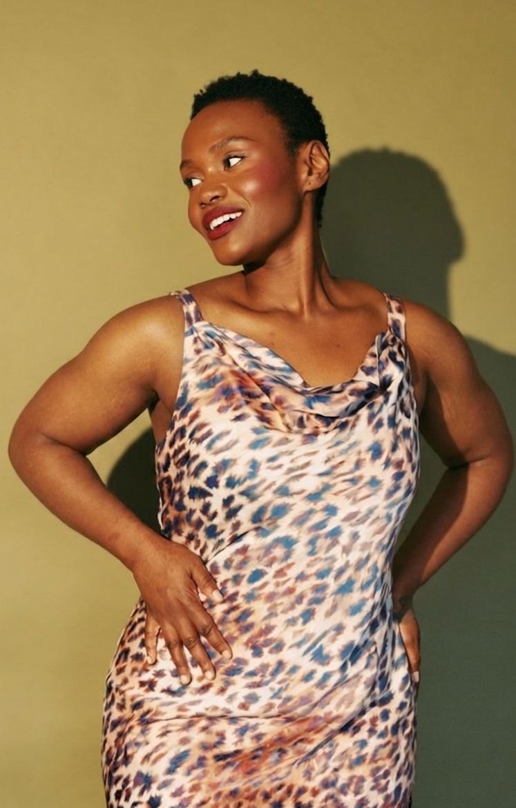 Hilary Biaku | Black Female Model, Creative Strategist, Content Creator, Public Speaker, and Life Coach — A smiling woman with short curly hair wearing a sleeveless animal print dress, posing with hands on hips against a yellow-green background.