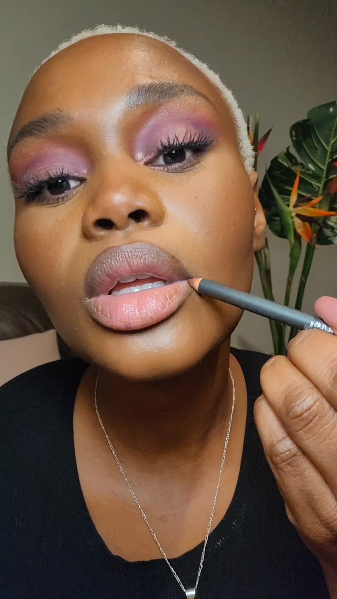 Hilary Biaku | Black Female Model, Creative Strategist, Content Creator, Public Speaker, and Life Coach —  A woman with short blonde hair and makeup applying lip color with a pencil in front of a leafy plant.