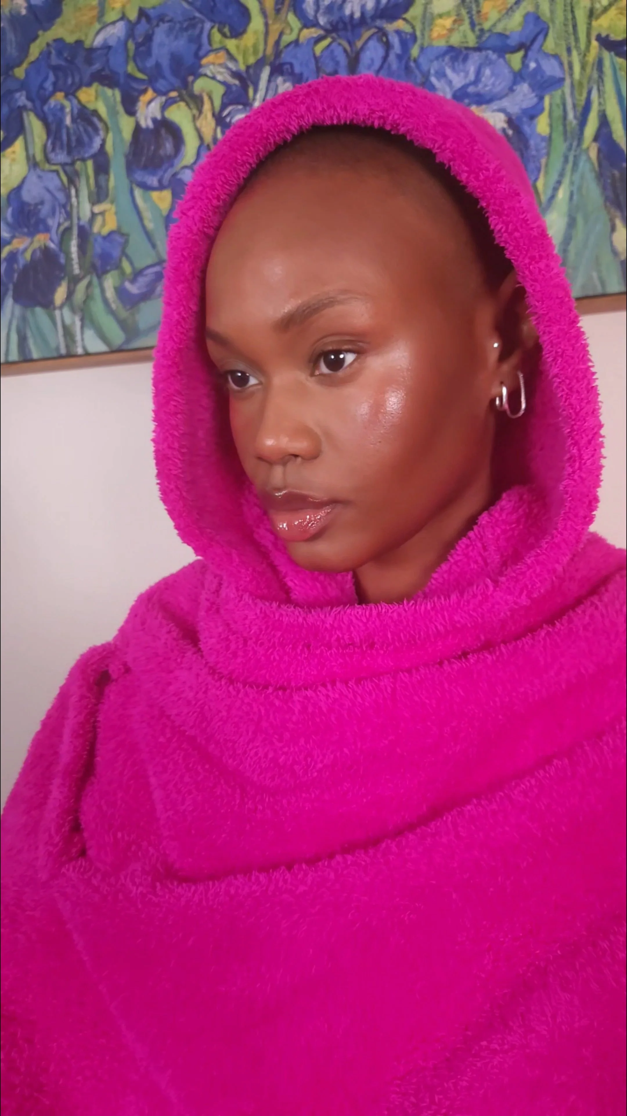 Hilary Biaku | Black Female Model, Creative Strategist, Content Creator, Public Speaker, and Life Close-up of a woman with a shaved head, wearing a bright pink fuzzy hoodie, standing in front of a wall with a painting of purple and yellow irises.