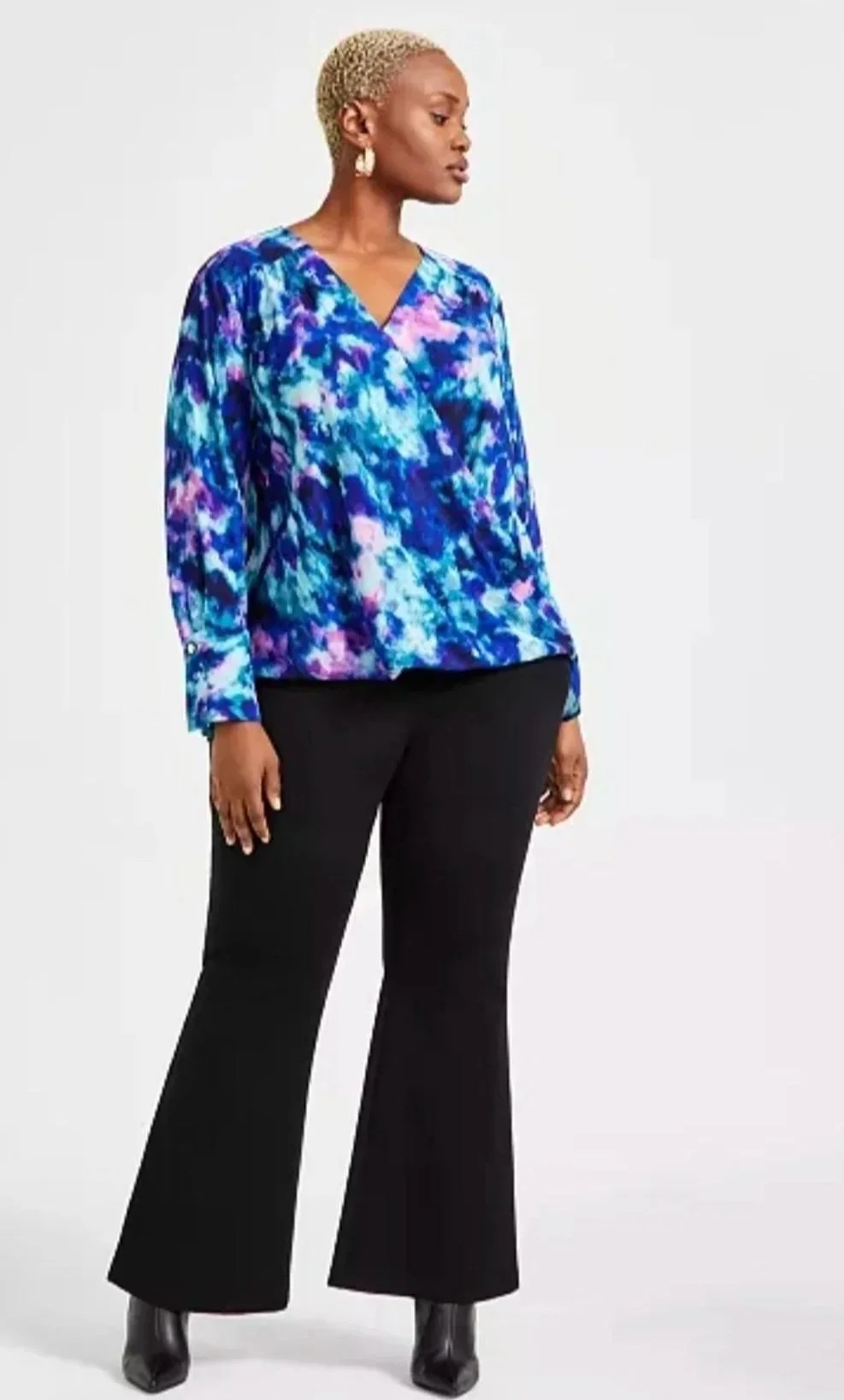 Hilary Biaku | Black Female Model, Creative Strategist, Content Creator, Public A woman with short blonde hair wearing a blue, purple, and pink abstract patterned blouse, black flared pants, and black boots, standing against a plain background.