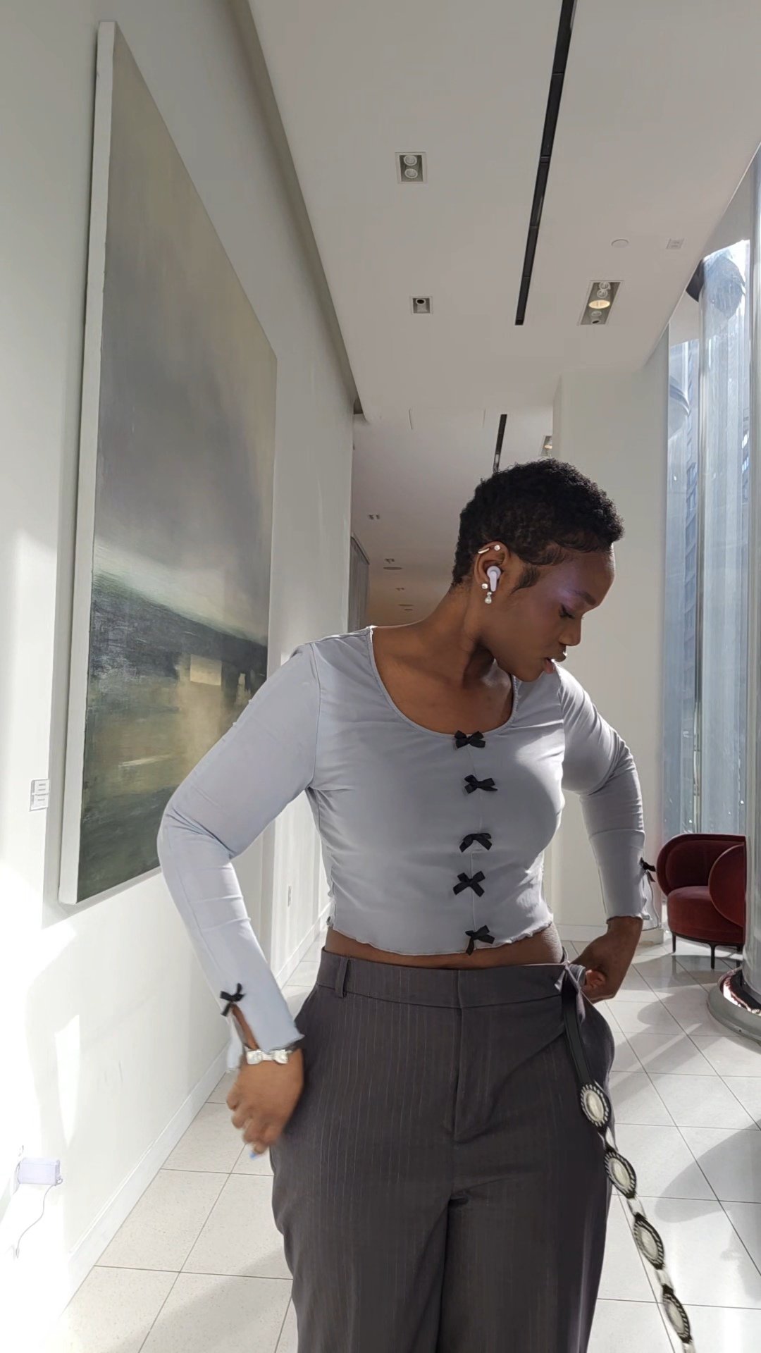 A woman with short hair dressed in a white top with black bow accents, gray pinstriped pants, and large earrings, standing indoors with her head lowered and hands on her hips.
