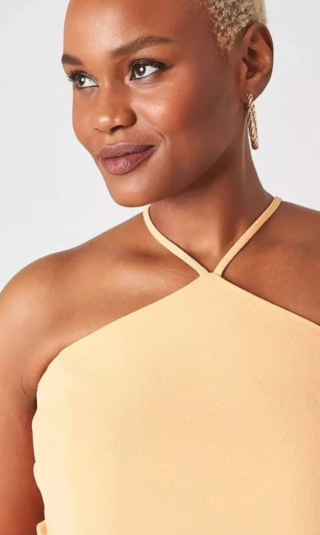 Hilary Biaku | Black Female Model, Creative Strategist, Content Creator, Public Speaker, and Life Coach — A close-up of a woman with short blonde hair, wearing gold hoop earrings and a beige halter top, smiling softly and looking to her right.