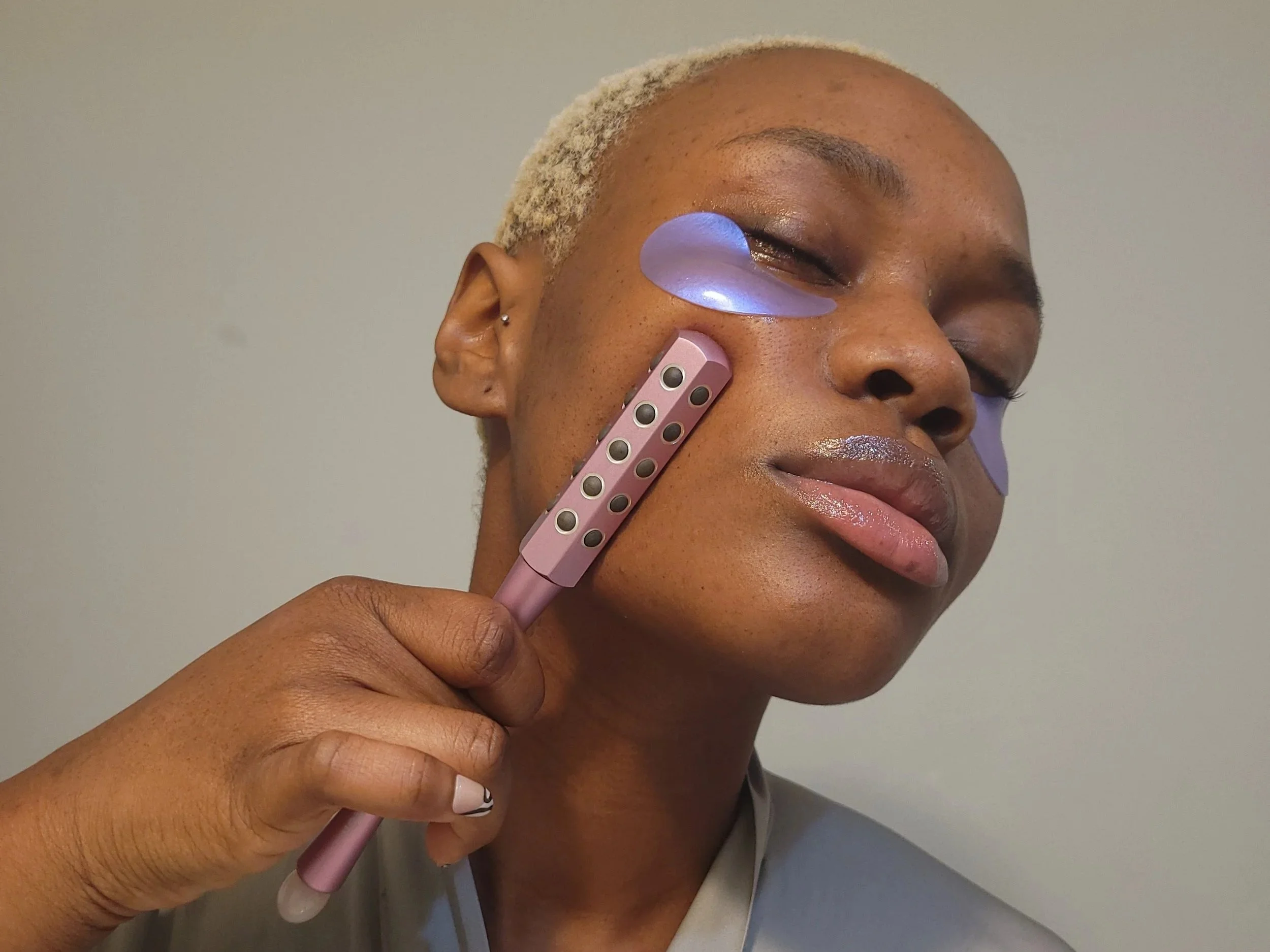 Hilary Biaku | Black Female Model, Creative Strategist, Content Creator, Public A person with short, blonde hair and dark skin holds a facial massager near their face with closed eyes, using a pink and purple device with multiple circular nodes.