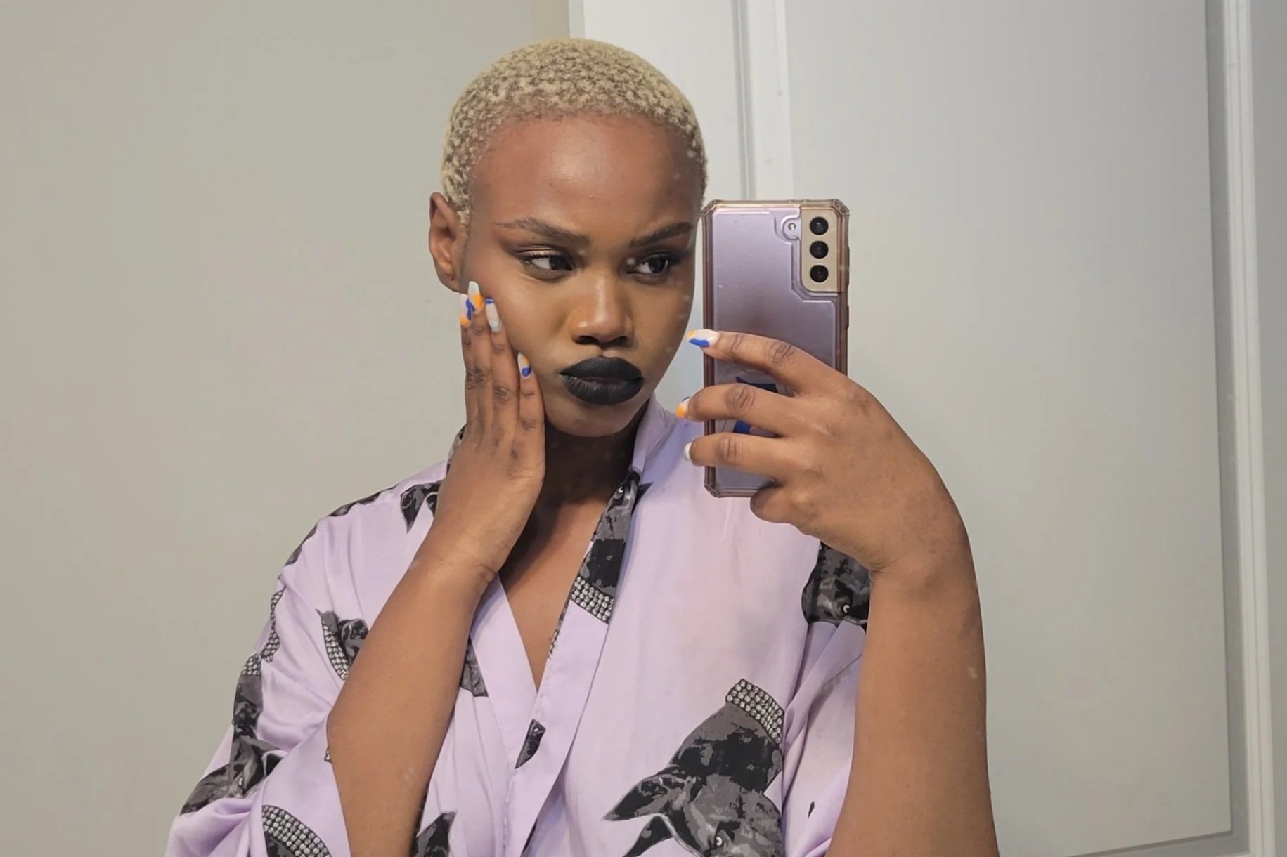 A woman with short blonde hair taking a mirror selfie with her smartphone. She is touching her face with one hand and wearing a lavender top with black and white patterned accents.
