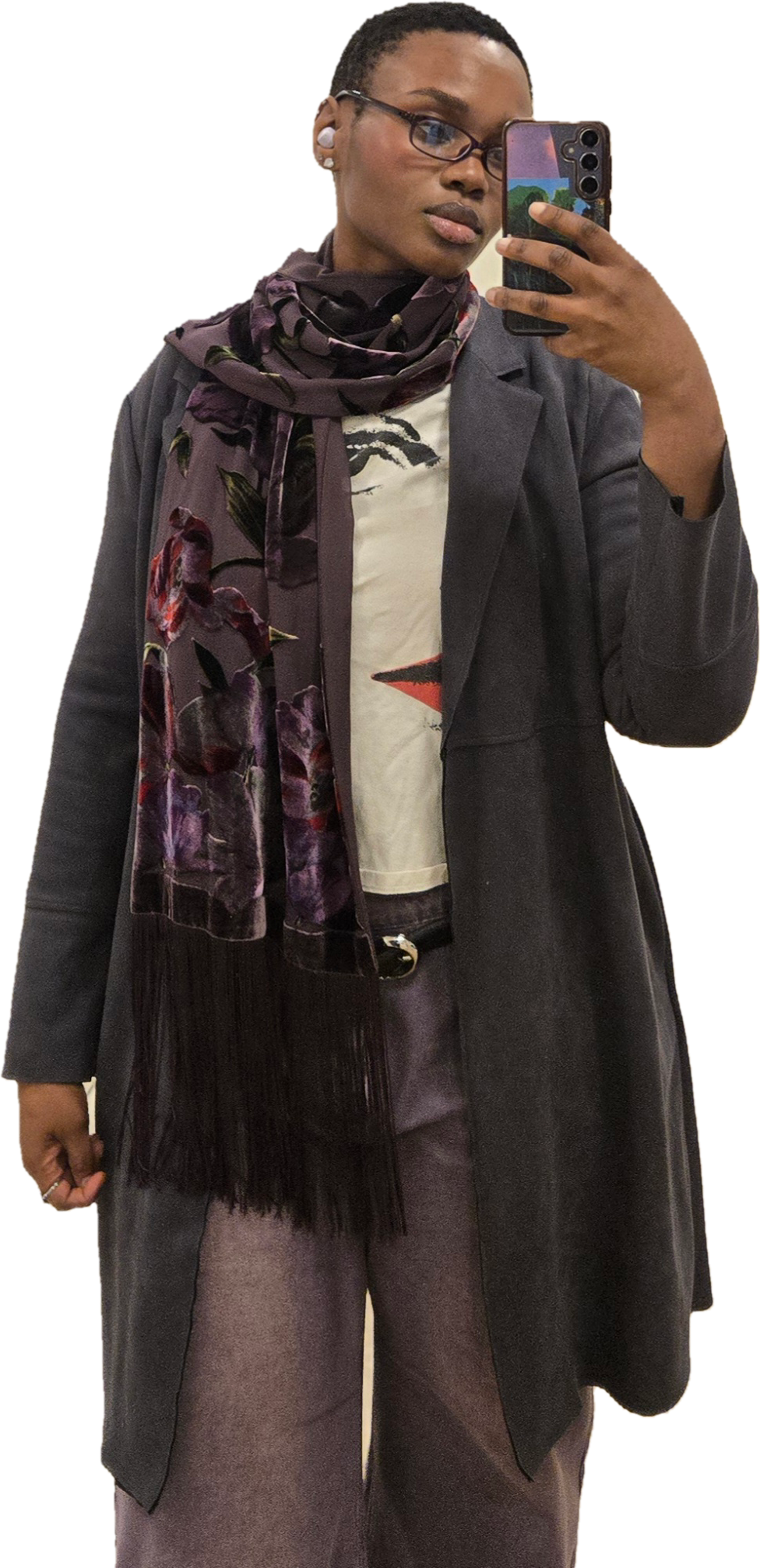 A woman taking a selfie with a smartphone, wearing glasses, a dark blazer, purple pants, a patterned shirt, and a floral scarf.