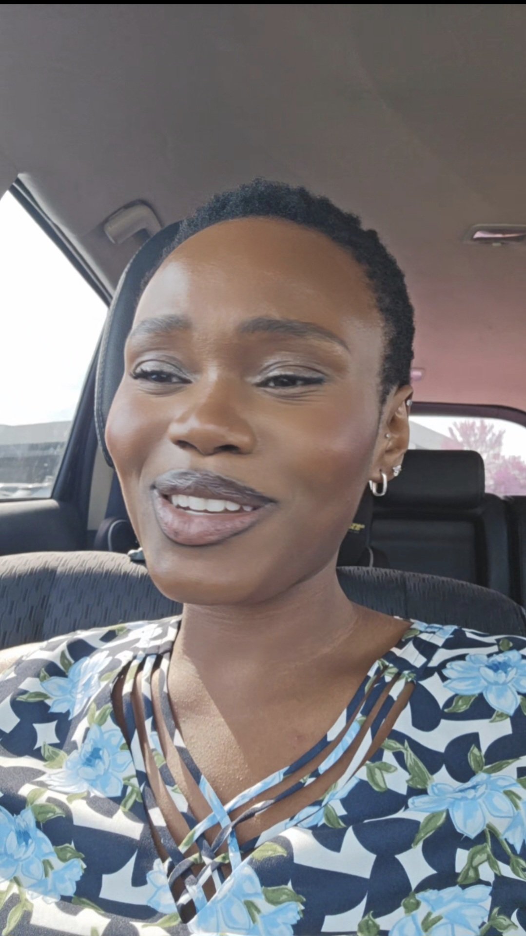 Hilary Biaku | Black Female Model, Creative Strategist, Content Creator, Public Speaker, and Life Coach — A woman with short curly hair smiling in a car, wearing a floral top with cut-out detail.
