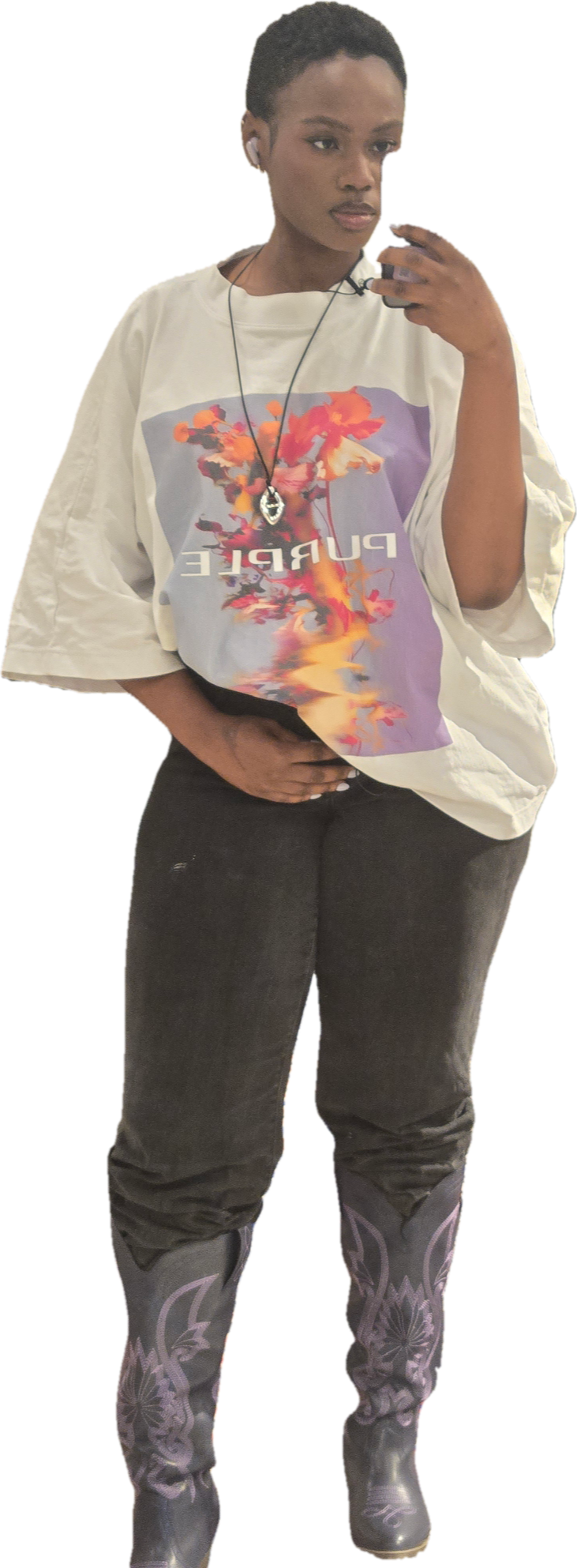 Hilary A woman taking a mirror selfie, wearing a white oversized graphic T-shirt with a colorful abstract design and the word 'PURPLE' on it, black pants, and patterned cowboy boots. She has short, natural hair, white earbuds, and a pendant necklace.