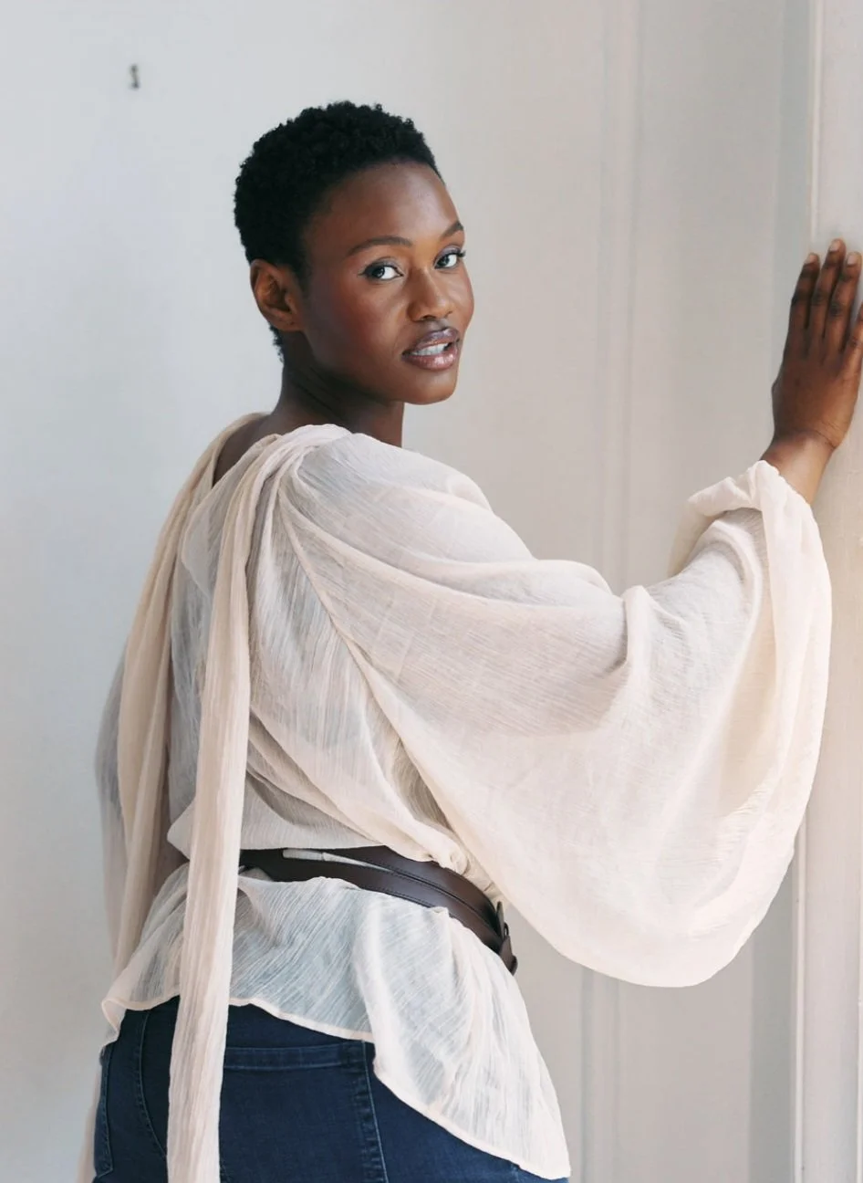Hilary Biaku | Black Female Model, Creative Strategist, Content Creator, Public Speaker, Black woman with short curly hair wearing a cream-colored, long-sleeved blouse and dark jeans, standing against a light-colored wall, looking over her shoulder.