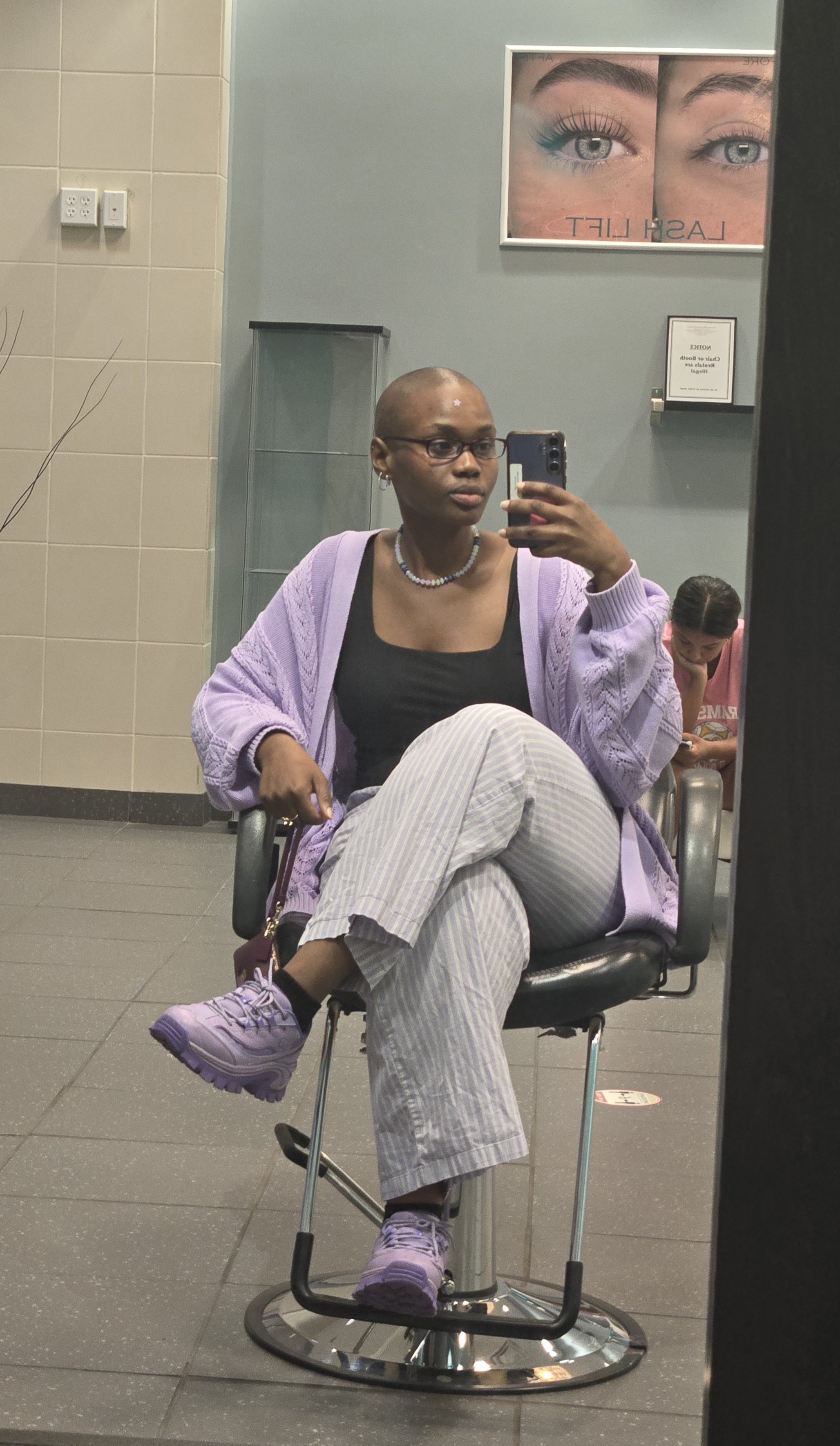 Woman sitting on a barber chair taking a selfie in a mirror, with a young girl in the background looking at her phone, in a barbershop or salon setting.